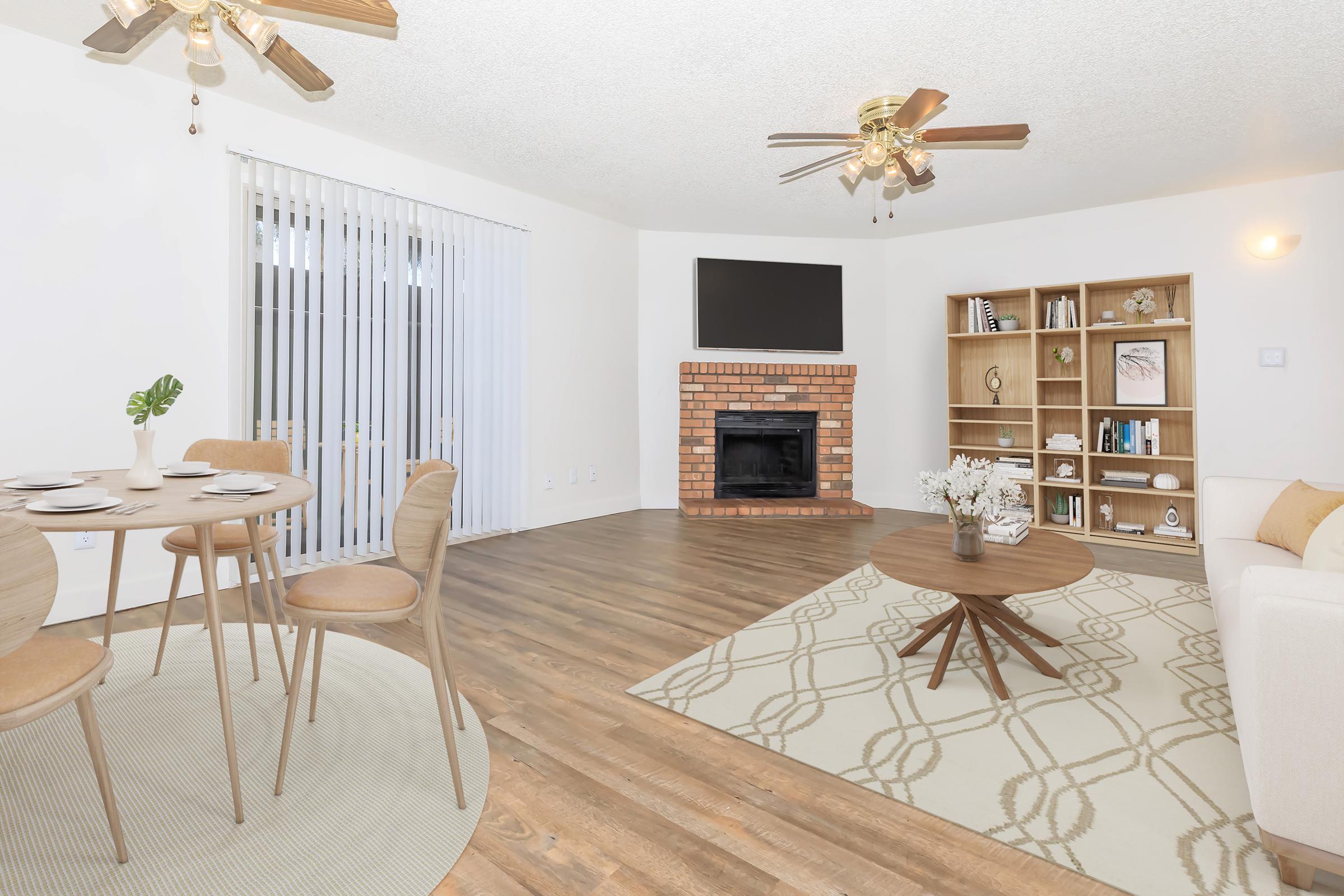 Cozy living space featuring a dining area with a round table and four chairs, a comfortable beige sofa, a coffee table, and a brick fireplace. The room has a modern aesthetic with wooden flooring, light-colored walls, and a TV mounted on the wall. Natural light filters through the blinds on sliding doors.