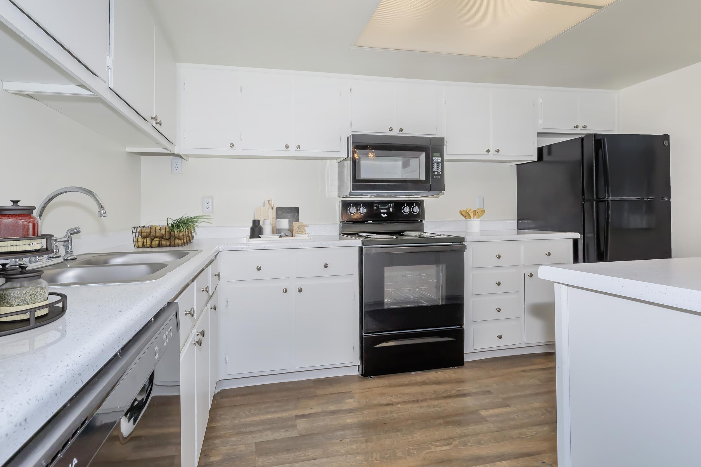 Modern kitchen featuring white cabinetry, a black stove and microwave, and a black refrigerator. Countertops are light-colored with a shiny finish. There is a sink with a dish rack and various kitchen utensils neatly arranged. The flooring is a wood-like laminate, creating a warm atmosphere.