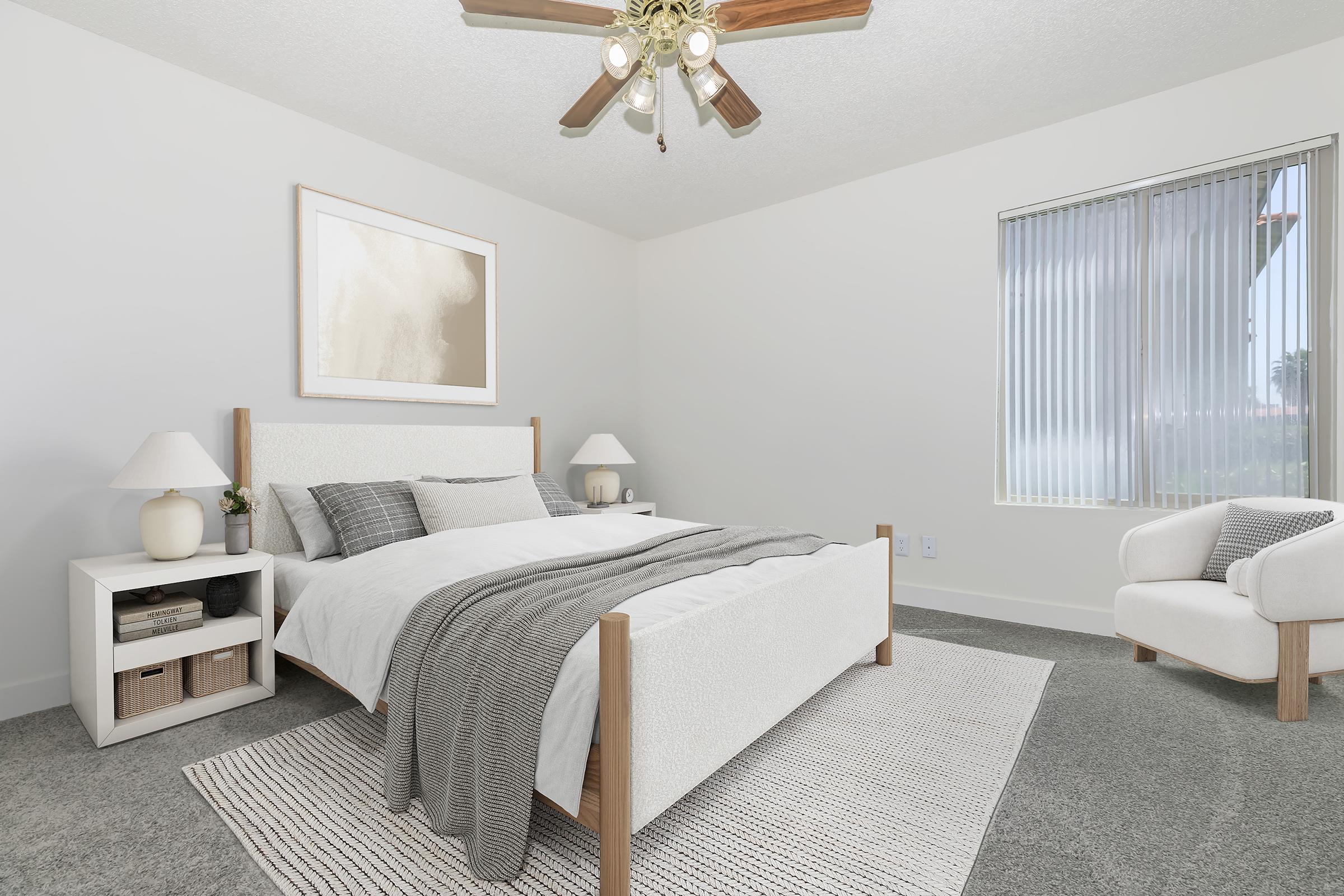 A modern bedroom featuring a light-colored bed with white bedding and a gray throw blanket. There are two matching nightstands with lamps, a decorative wall art piece, and a cozy armchair. The room has gray carpet and large windows with blinds, allowing natural light to fill the space.