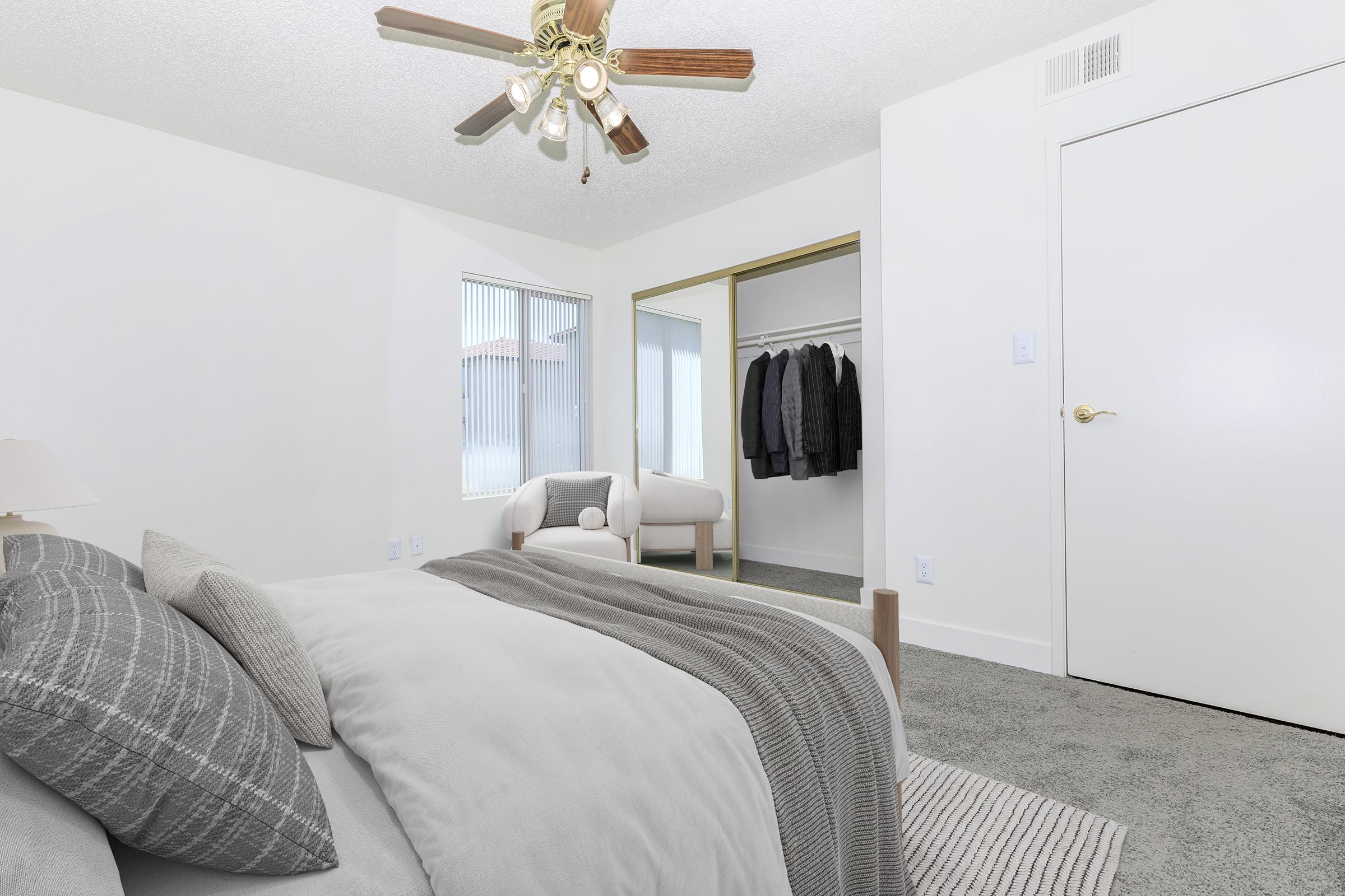A well-lit, modern bedroom featuring a cozy bed with decorative pillows, a ceiling fan, and a light-colored carpet. In the background, there's a mirrored closet with clothing hanging, and a small seating area with two chairs. The walls are painted white, creating a fresh and inviting atmosphere.