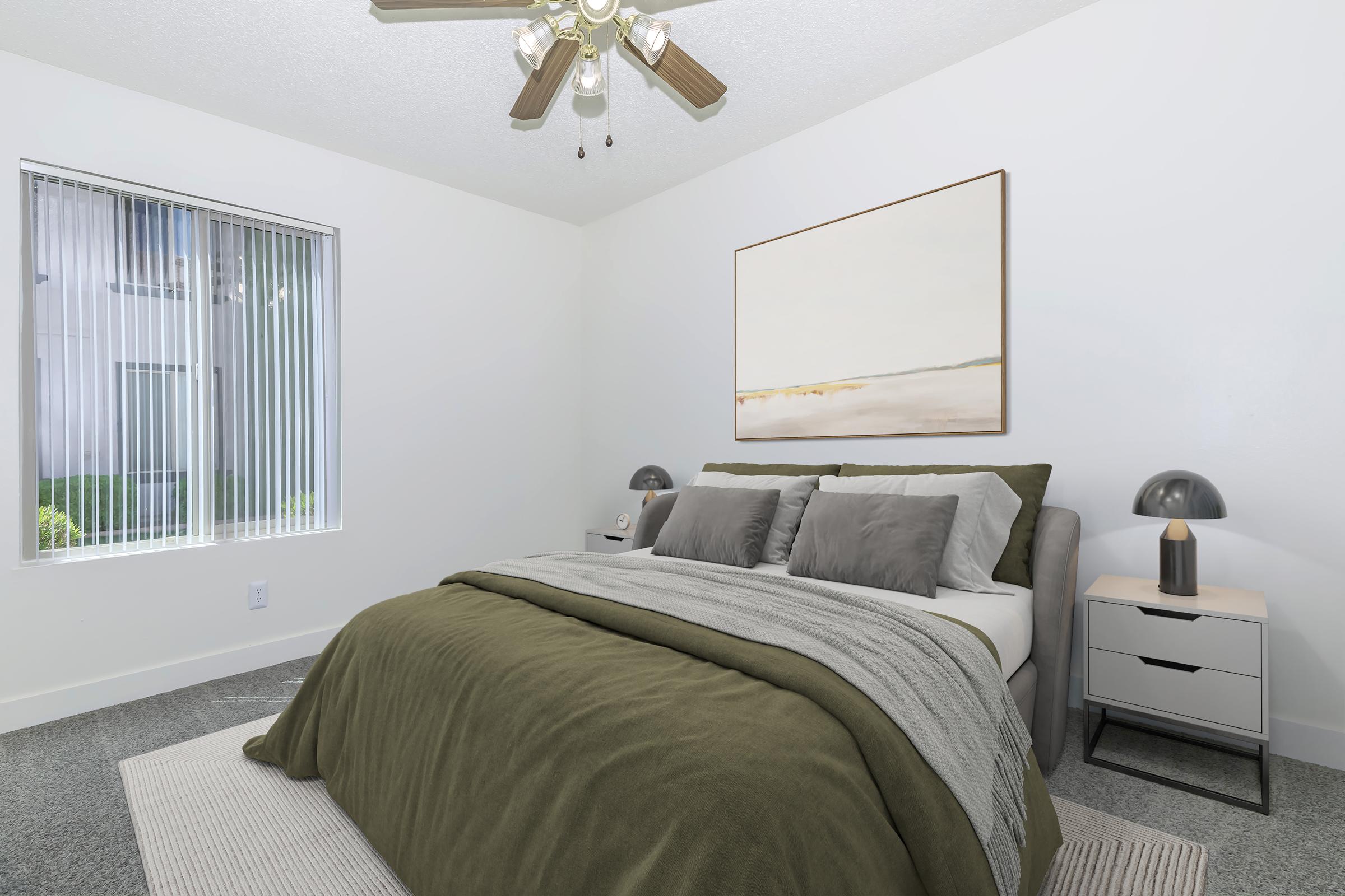 A modern bedroom featuring a queen-sized bed with gray and green bedding, a bedside table with a lamp, and a large piece of abstract artwork on the wall. The room has a ceiling fan and a window with vertical blinds, allowing natural light to fill the space. A light-colored area rug adds warmth to the floor.