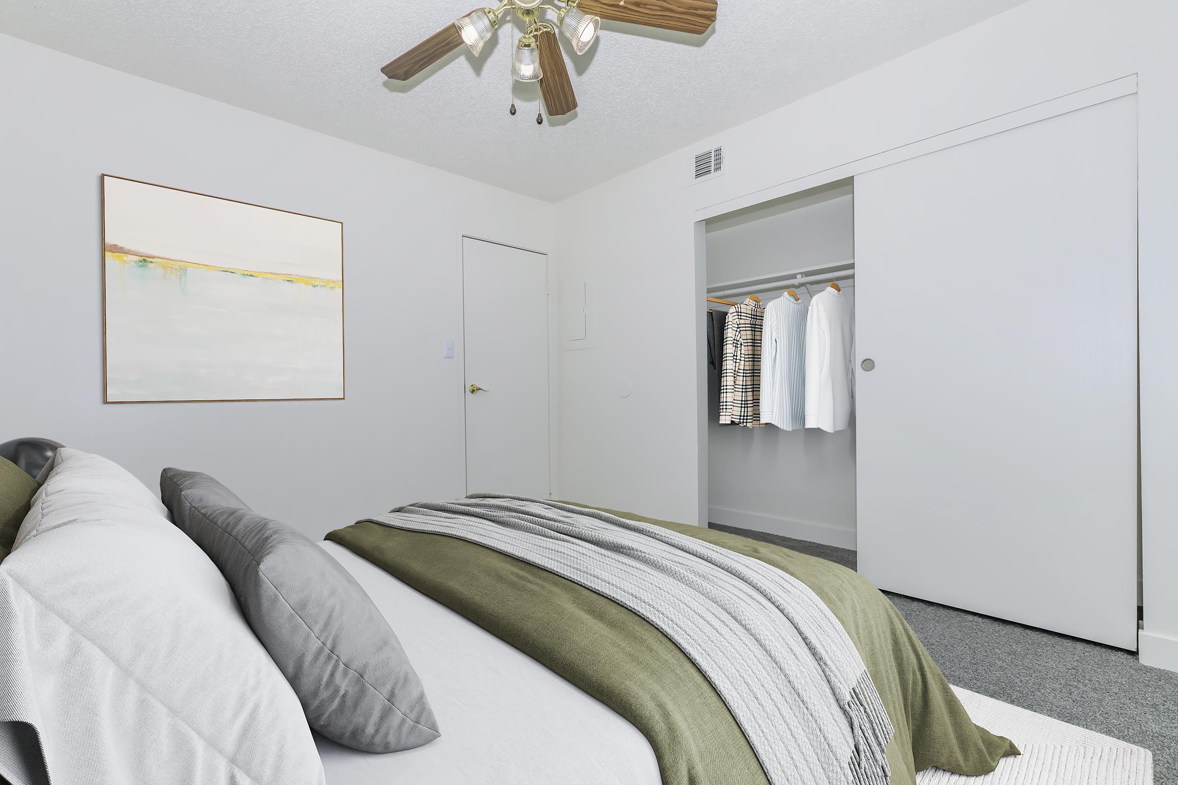 A cozy, modern bedroom featuring a neatly made bed with gray and green bedding. A ceiling fan hangs overhead, and a piece of abstract art adorns the wall. A closet with sliding doors is visible, showcasing several hanging white shirts. The room has light-colored walls and carpet.