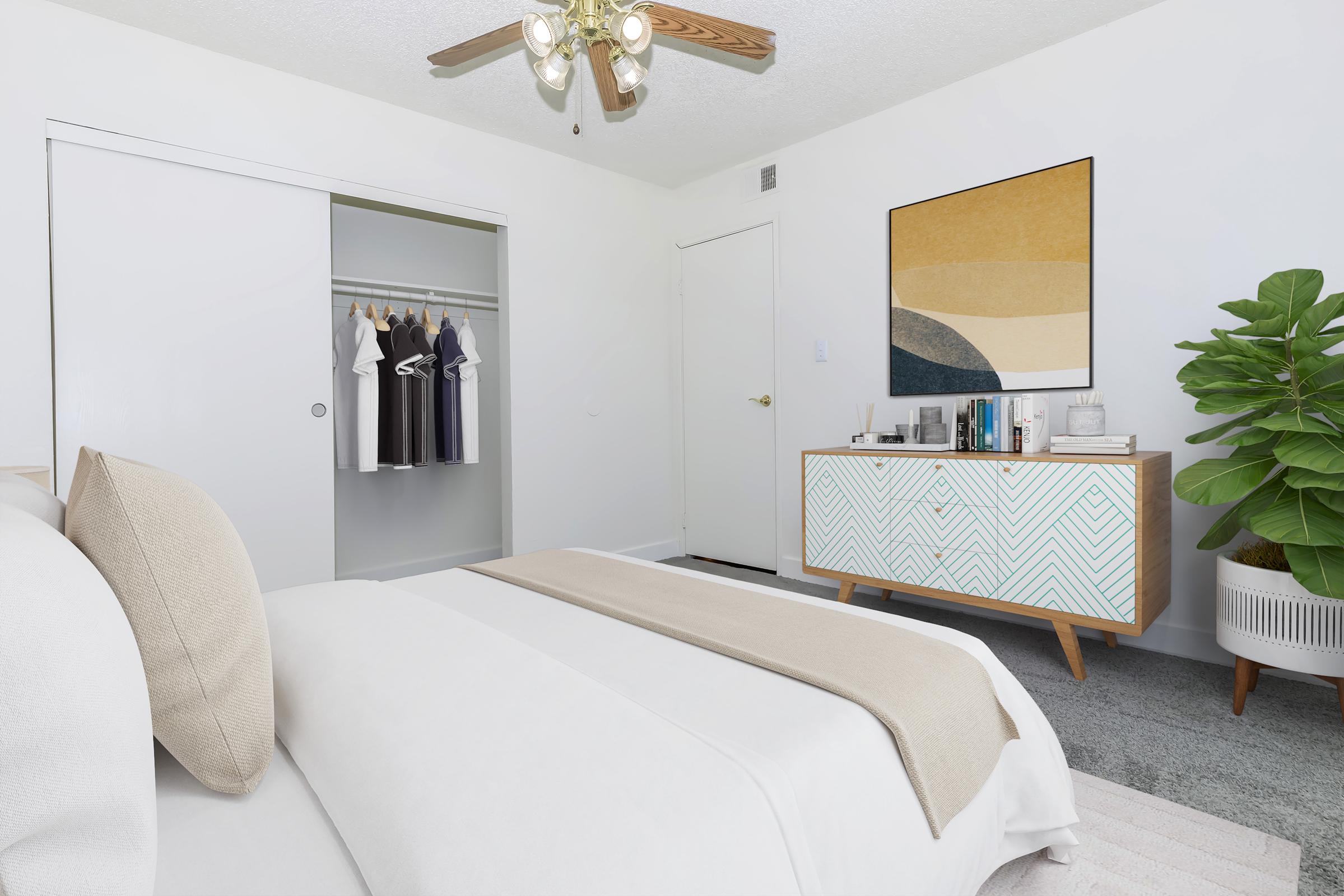 A modern bedroom featuring a neatly made bed with a beige throw blanket, a stylish side table with decorative items, and a dresser with geometric patterns. A closet is seen in the background with neatly hung clothes, and a large potted plant adds a touch of greenery to the room. Bright, neutral colors dominate the space.