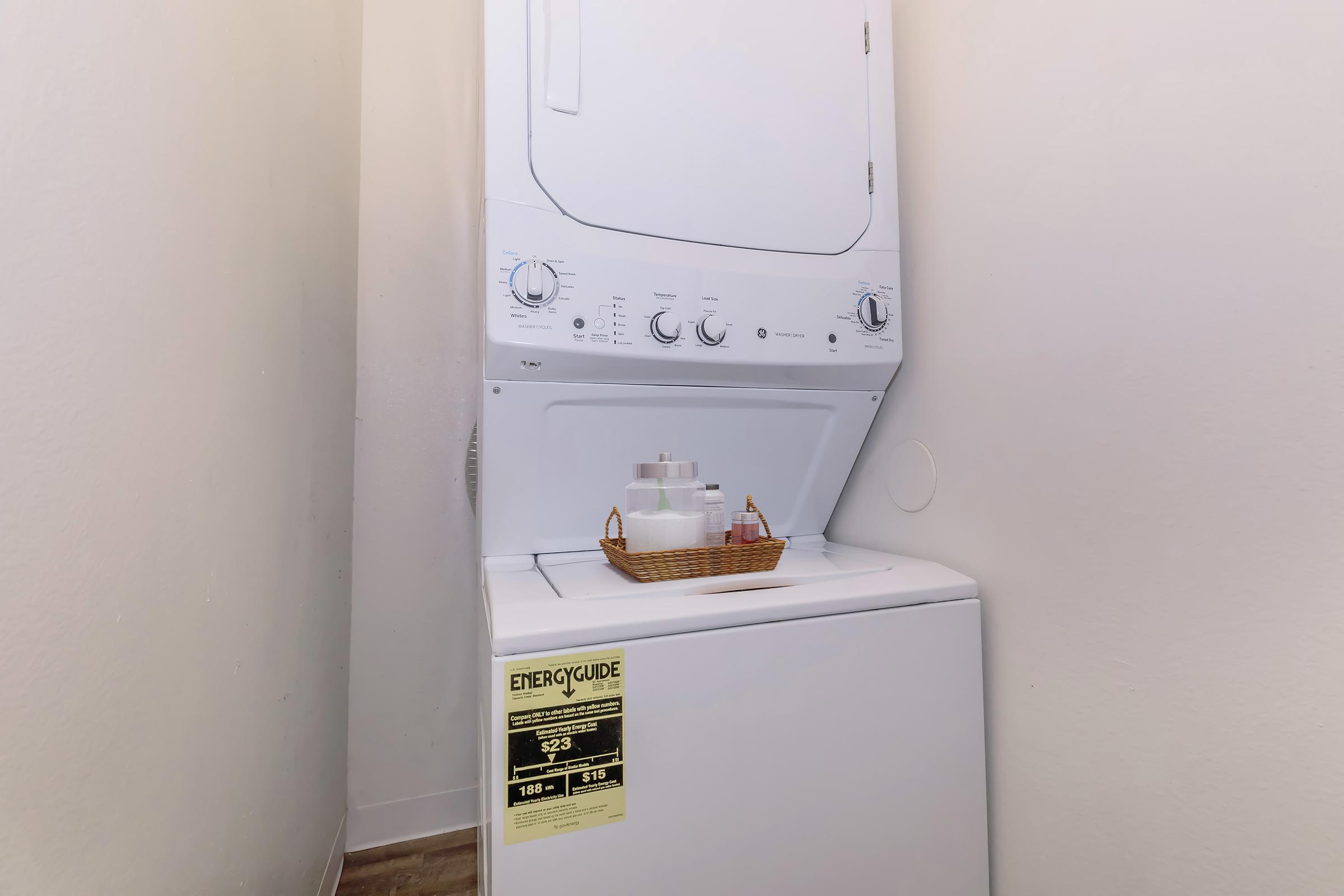 A stacked washer and dryer unit located in a small laundry space, accompanied by a basket containing laundry supplies. The unit has control knobs and an energy guide label visible on the front. The walls are plain, and the flooring is simple and clean.