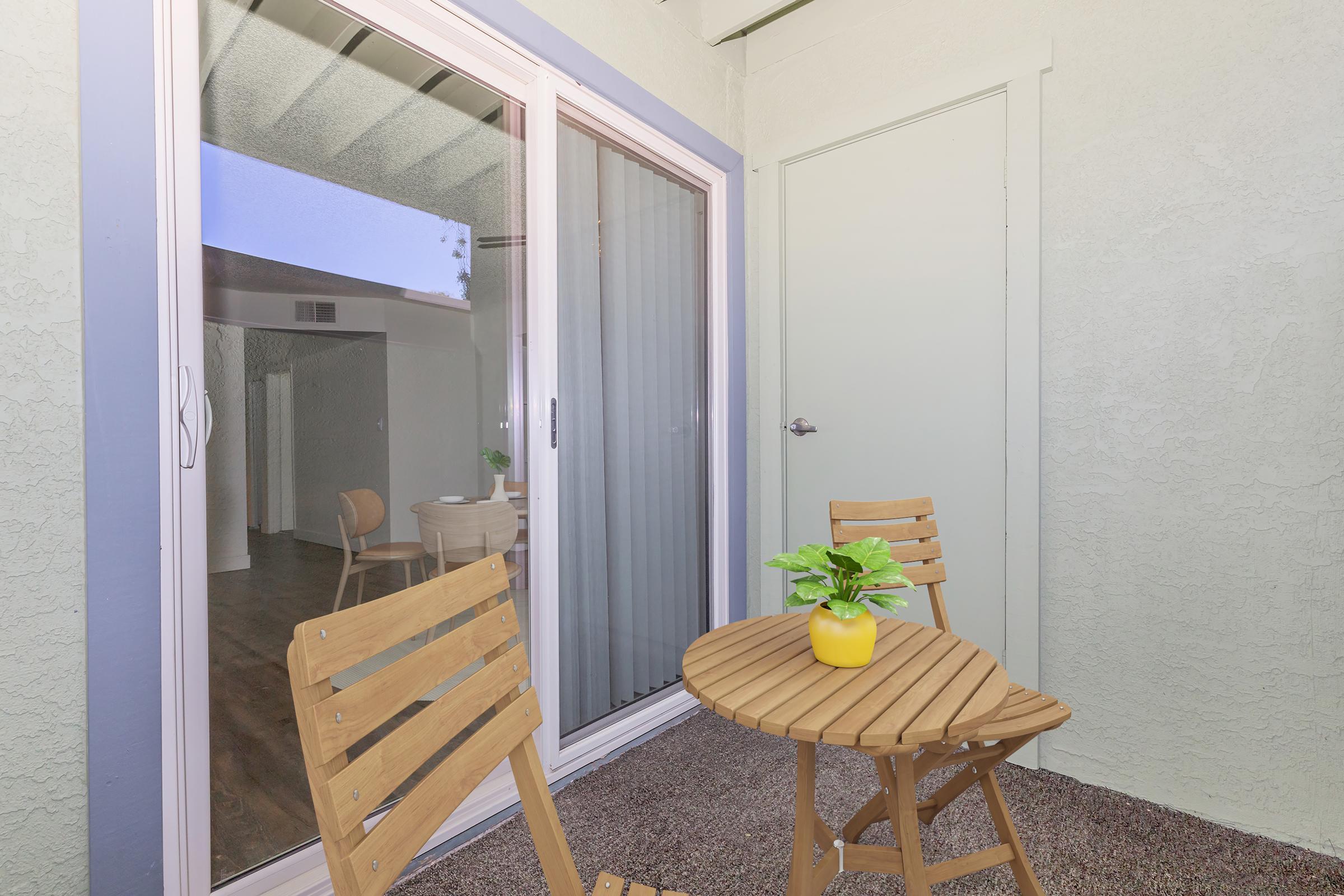 A small outdoor balcony area with a round wooden table and two matching chairs. A yellow plant pot with a green plant sits on the table. Sliding glass doors lead to an indoor space, with light brown flooring visible through the door. The walls are painted in light colors, giving a fresh and inviting look.