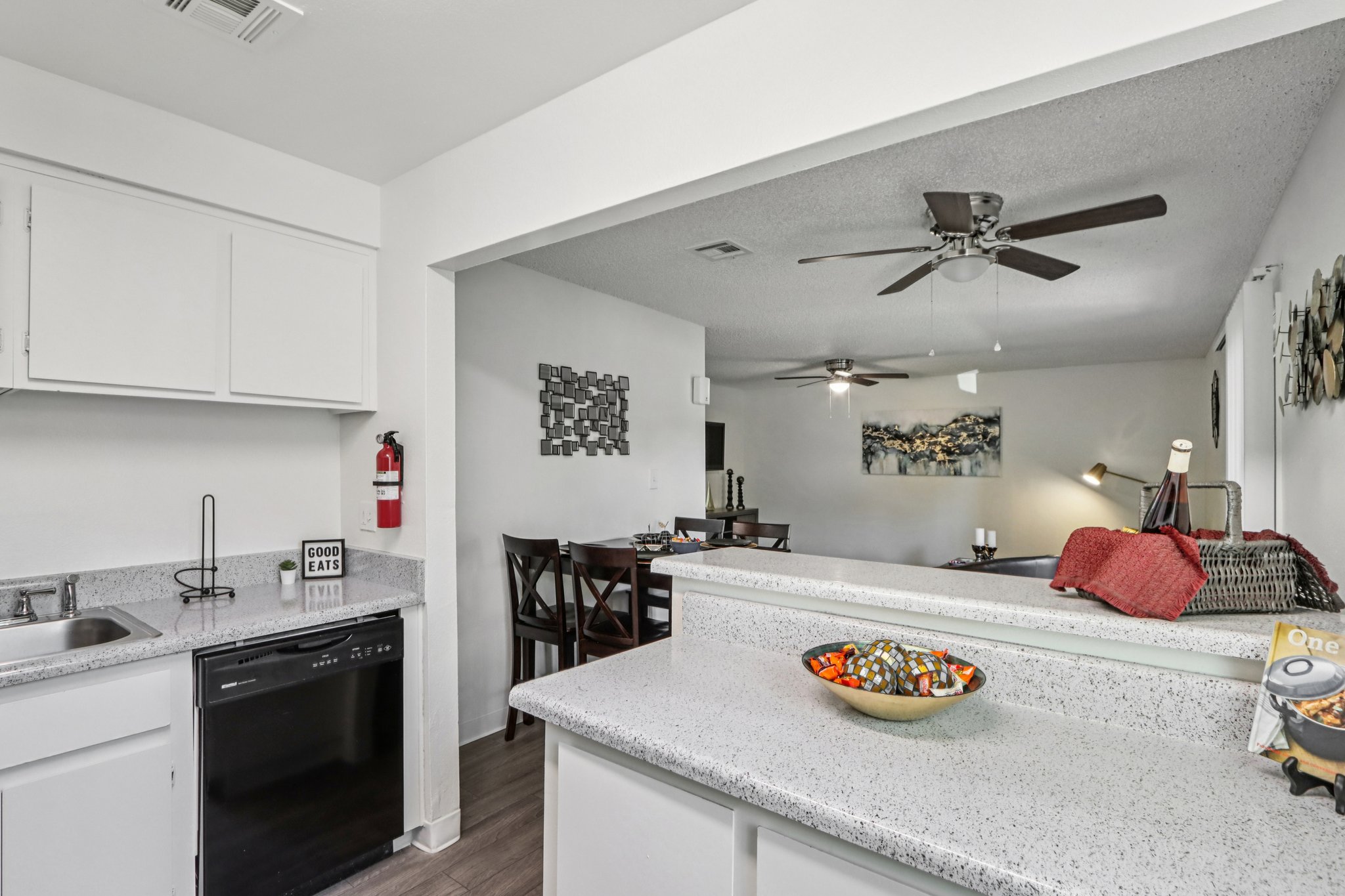 Modern kitchen interior featuring a gray countertop, black dishwasher, and a decorative bowl of fruit. The adjacent dining area has a wooden dining table and chairs, with a living space visible in the background, including a ceiling fan and artwork on the walls.