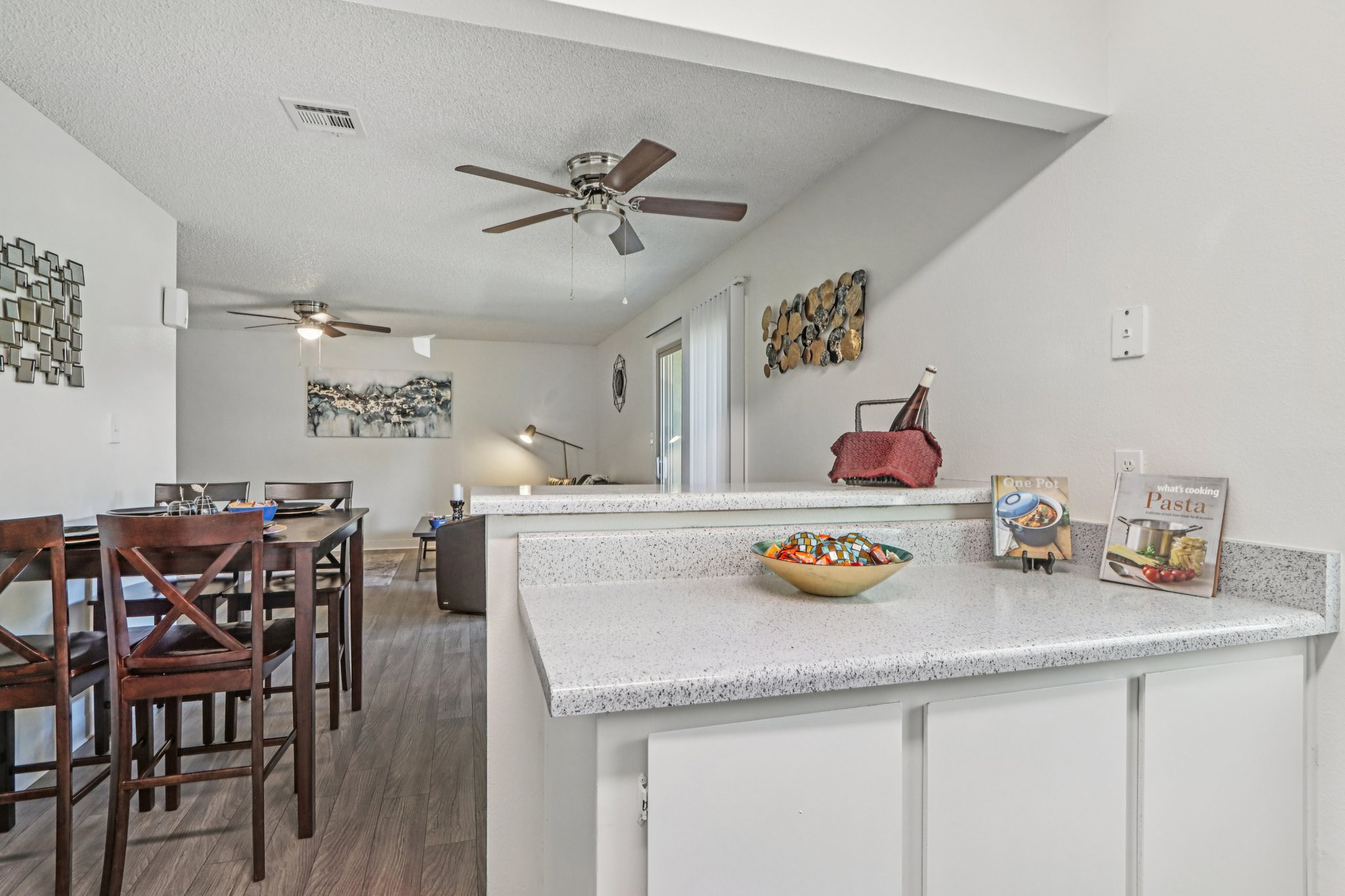 A view of a modern living space featuring a kitchen bar with a decorative bowl of fruit, dining area with wooden chairs, and a cozy seating area. There are two ceiling fans and artwork on the walls, creating a warm and inviting atmosphere. Natural light fills the room from an adjacent window.