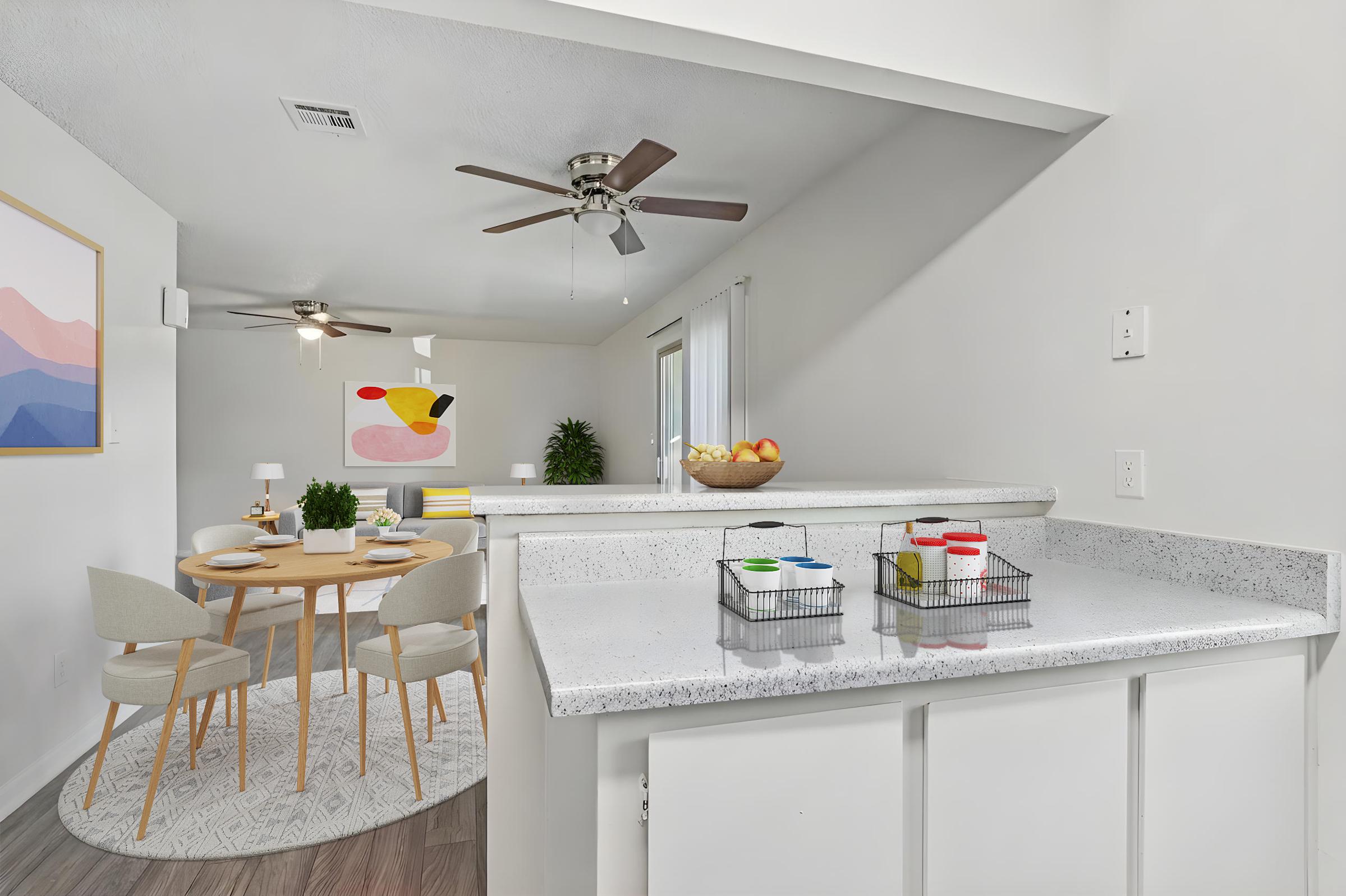 A modern kitchen with a granite countertop and colorful storage bins. In the background, there's a dining area featuring a round table with light chairs, a patterned rug, and a colorful wall art piece. Fresh fruit sits in a bowl on the countertop, and plants add a touch of greenery to the space.