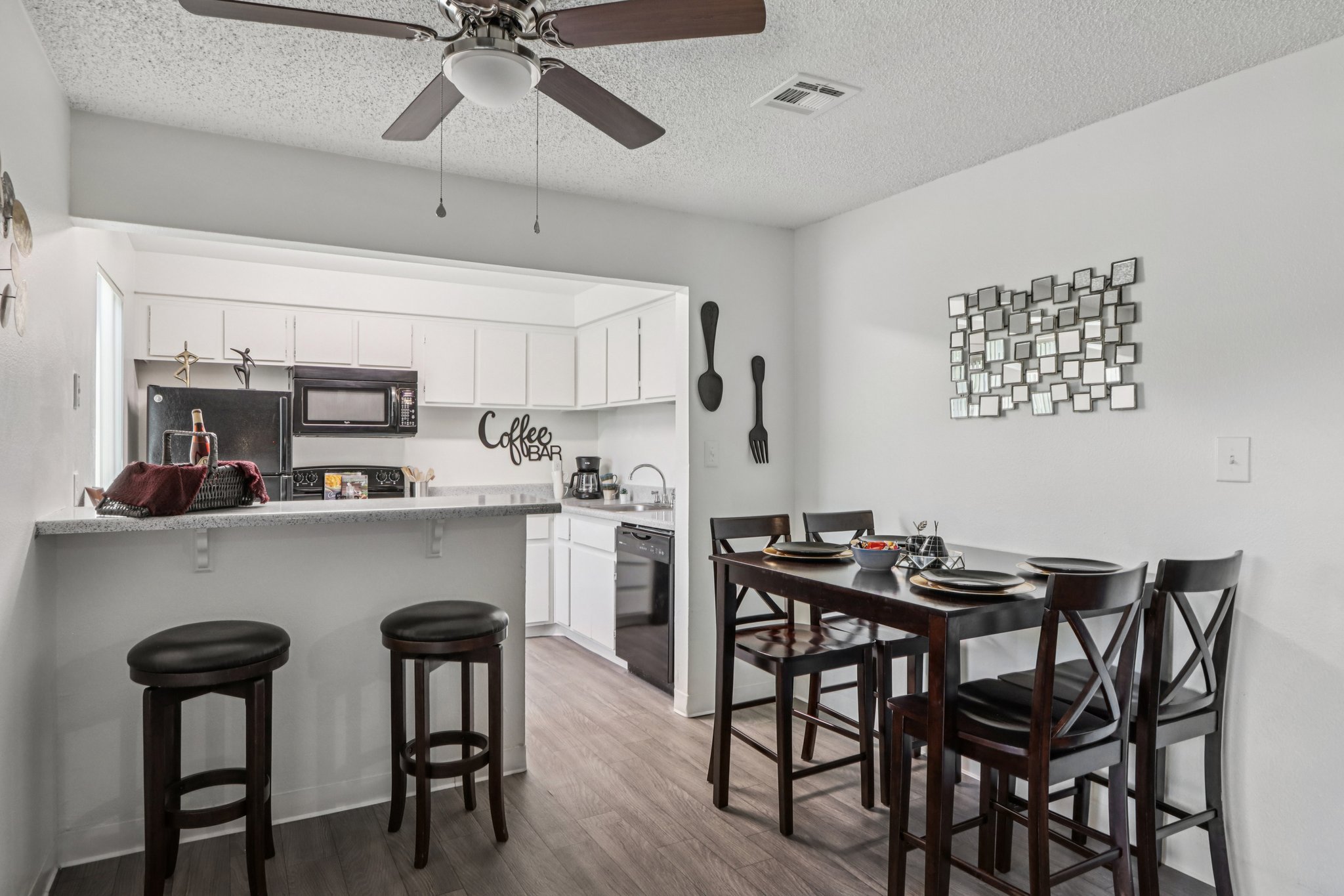 A modern kitchen and dining area featuring a small table with four black chairs, two bar stools at a breakfast bar, and stainless steel appliances. The kitchen has white cabinetry, a coffee decor piece on the wall, and a stylish wall mirror reflecting the space. The flooring is a light wood finish.