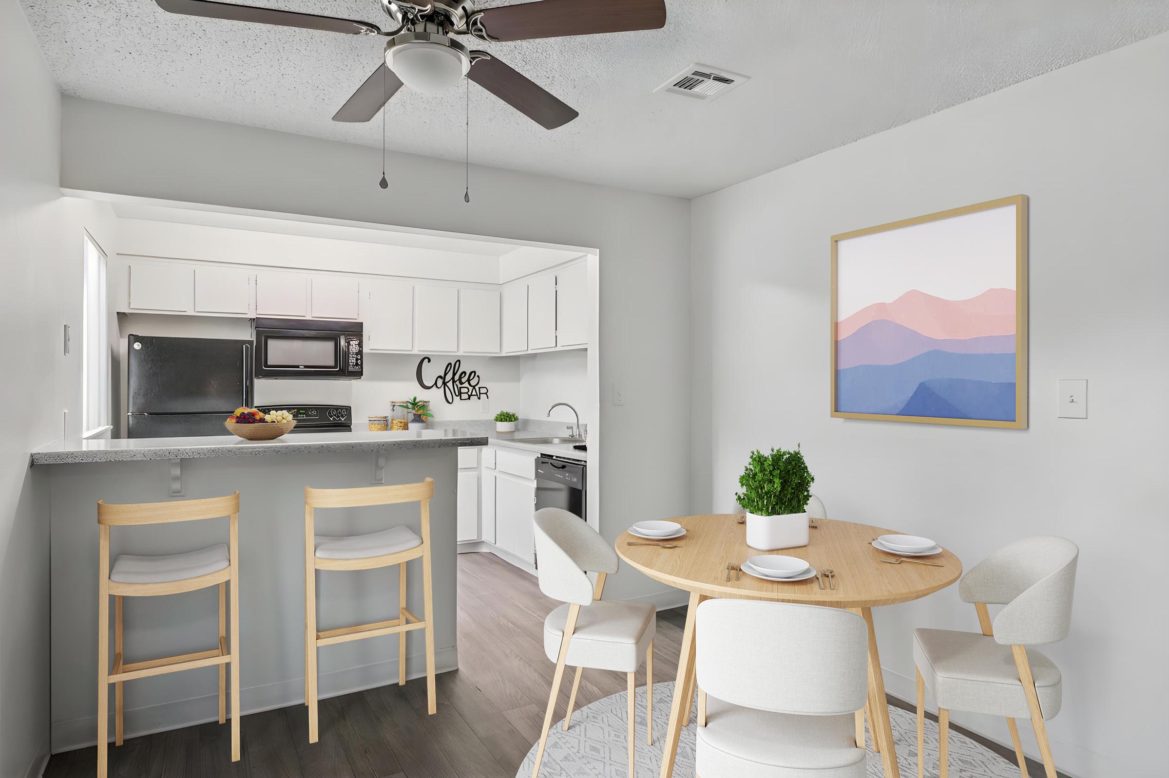 A modern kitchen and dining area featuring a round wooden table with four chairs, a ceiling fan, and an abstract painting on the wall. The kitchen, visible in the background, has white cabinets, a sink, and appliances, while a bowl of fruit is placed on the table in the foreground.