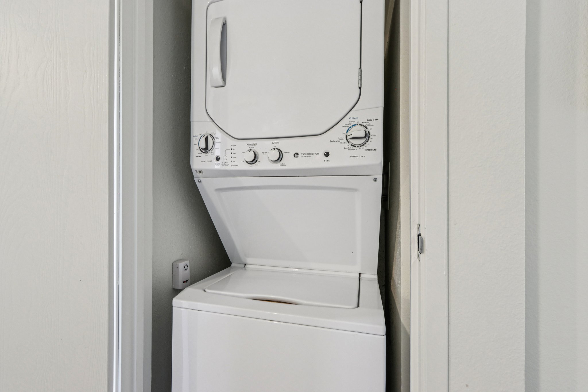 A stacked washer and dryer unit located in a narrow space between two walls. The washing machine is on the bottom and the dryer is on top, featuring dials and buttons for operation. The surrounding walls are painted in a light color, creating a clean, organized laundry area.