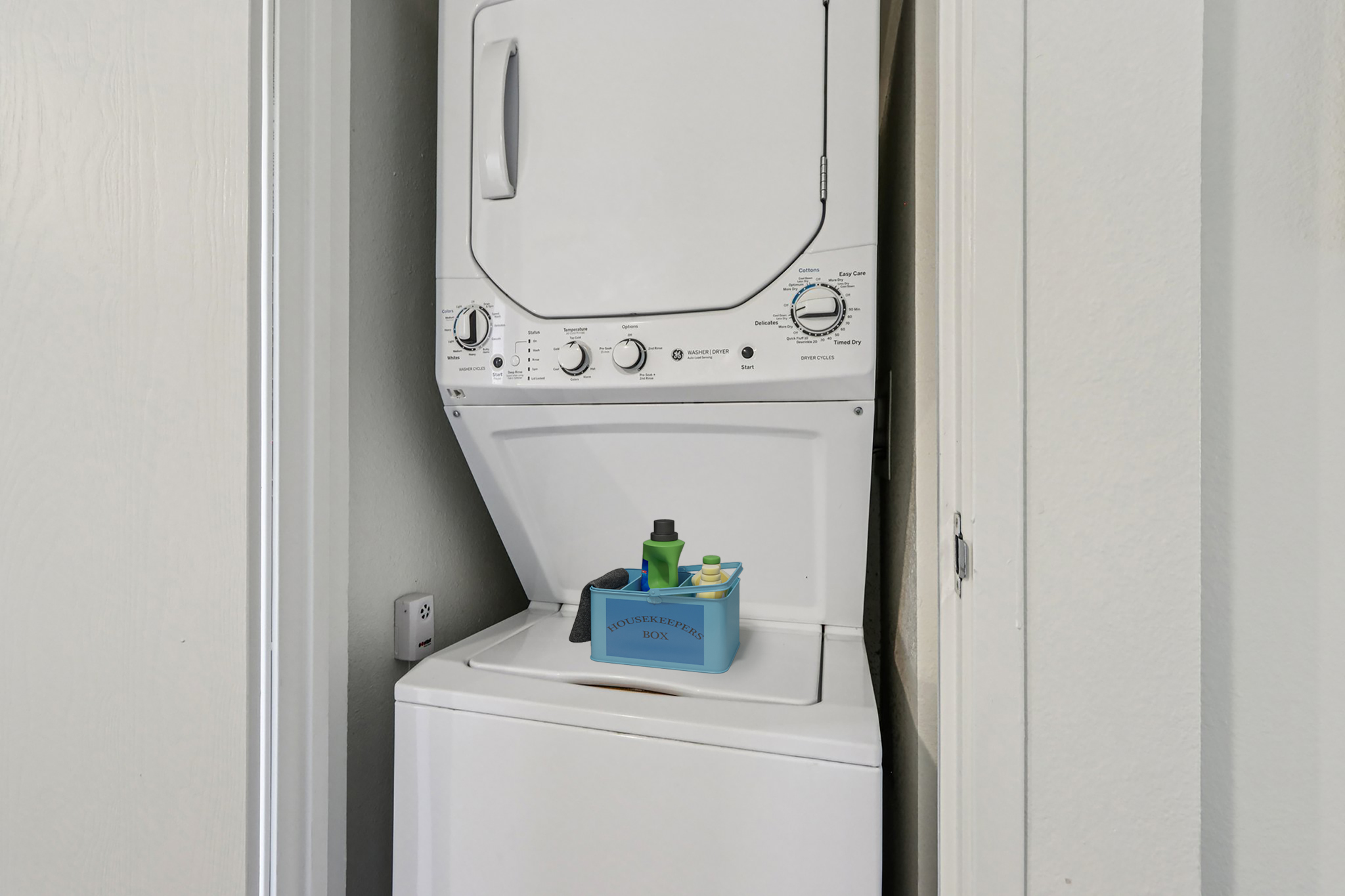 A stacked washer and dryer set in a laundry area, with a blue container labeled "Laundry Basket" sitting on top of the washing machine. The walls are light-colored, and the space appears clean and organized.