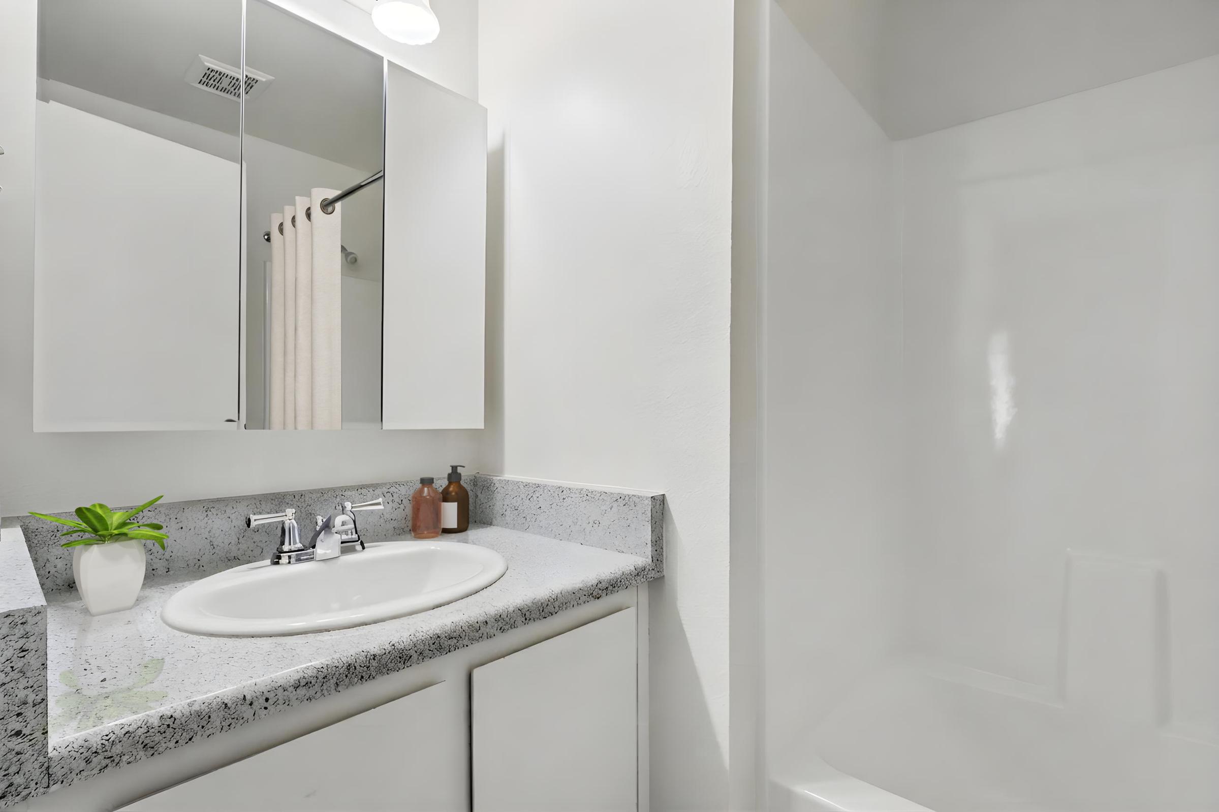 A clean, modern bathroom featuring a white countertop with a round sink, a small potted plant, and two bottles. Above the sink, there is a mirrored cabinet. A light-colored shower area is on the right, with a simple shower stall and light-colored walls, providing a bright and spacious appearance.