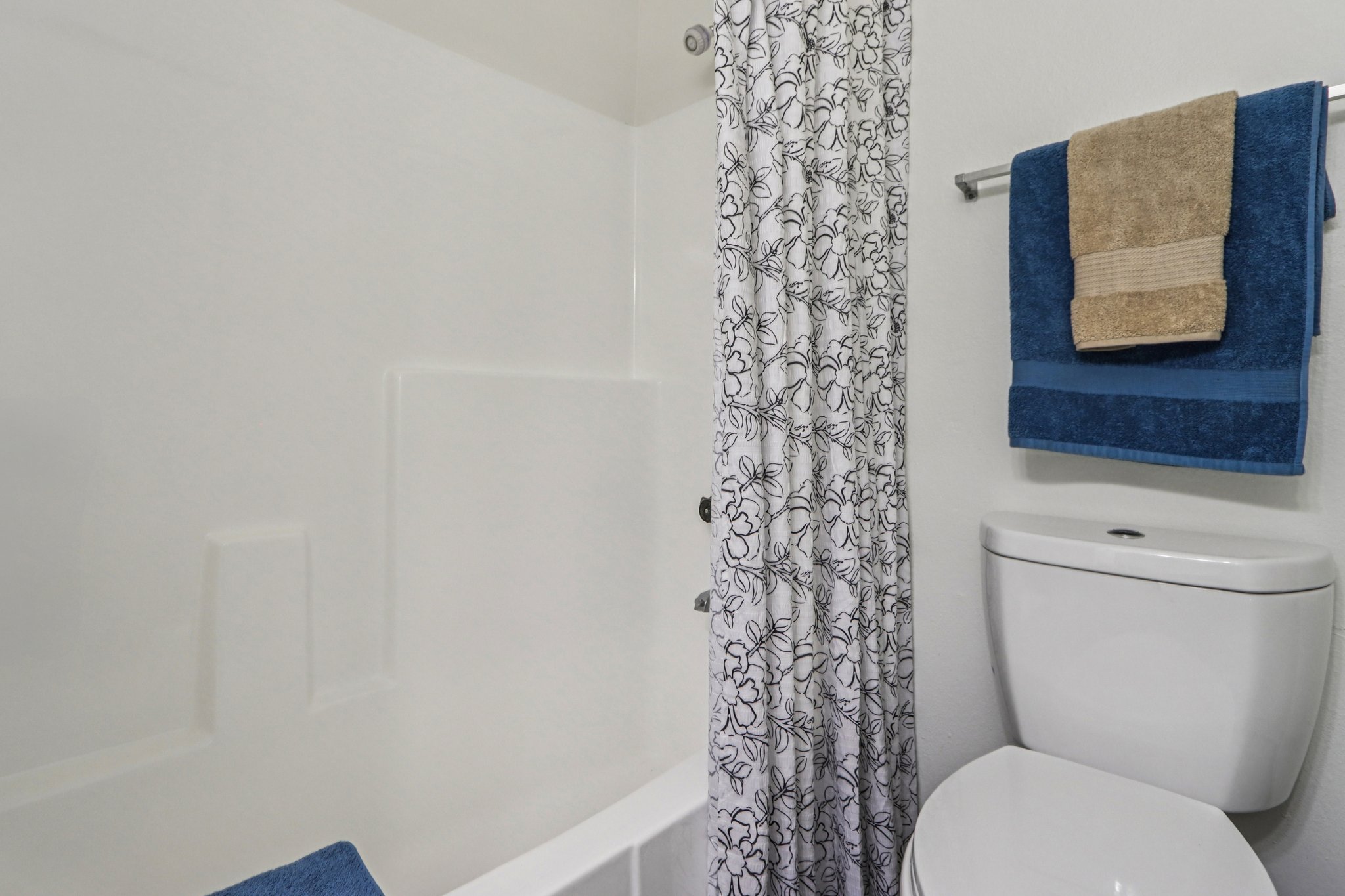 A clean bathroom featuring a white bathtub with a floral shower curtain, a toilet, and neatly arranged towels in blue and beige on a towel rack. The walls are light-colored, creating a bright and tidy atmosphere.