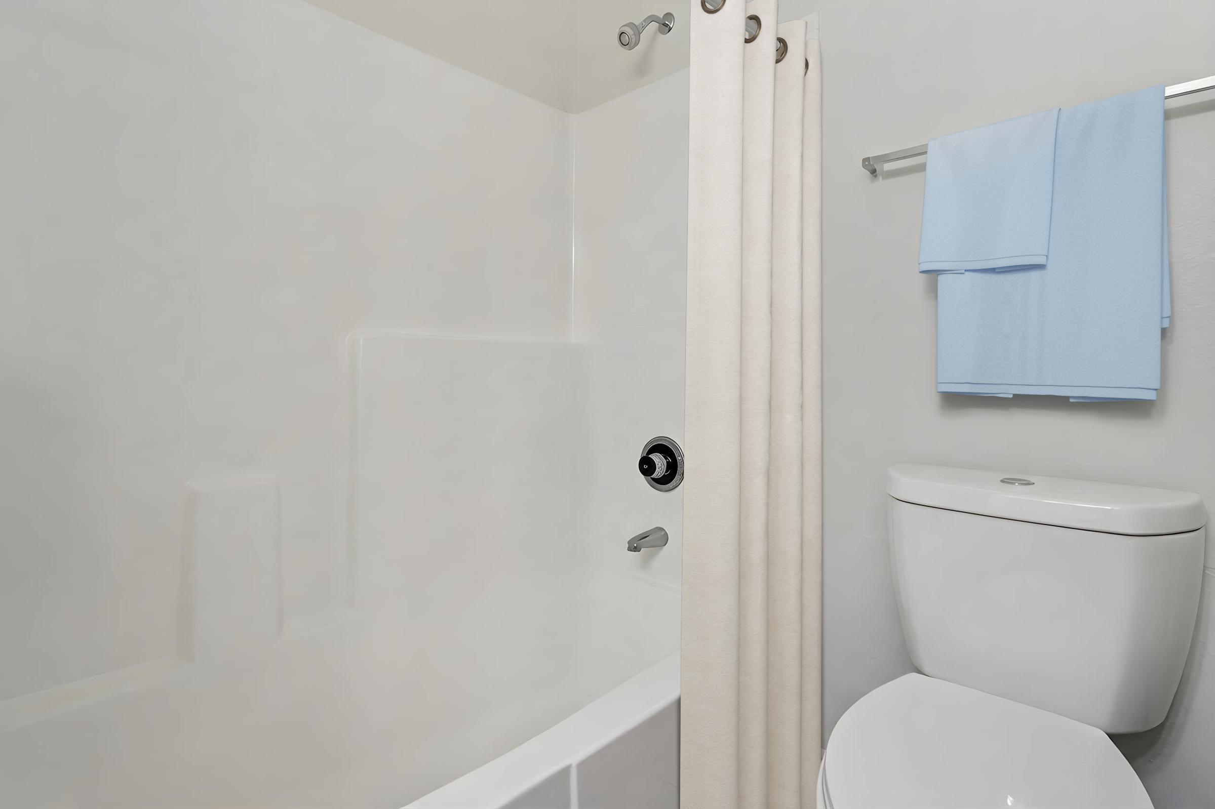 A modern bathroom showing a bathtub with a shower curtain, a toilet, and a towel rack holding two light blue towels. The walls are plain white, giving a clean and minimalist appearance. Natural lighting emphasizes the bright and airy feel of the space.