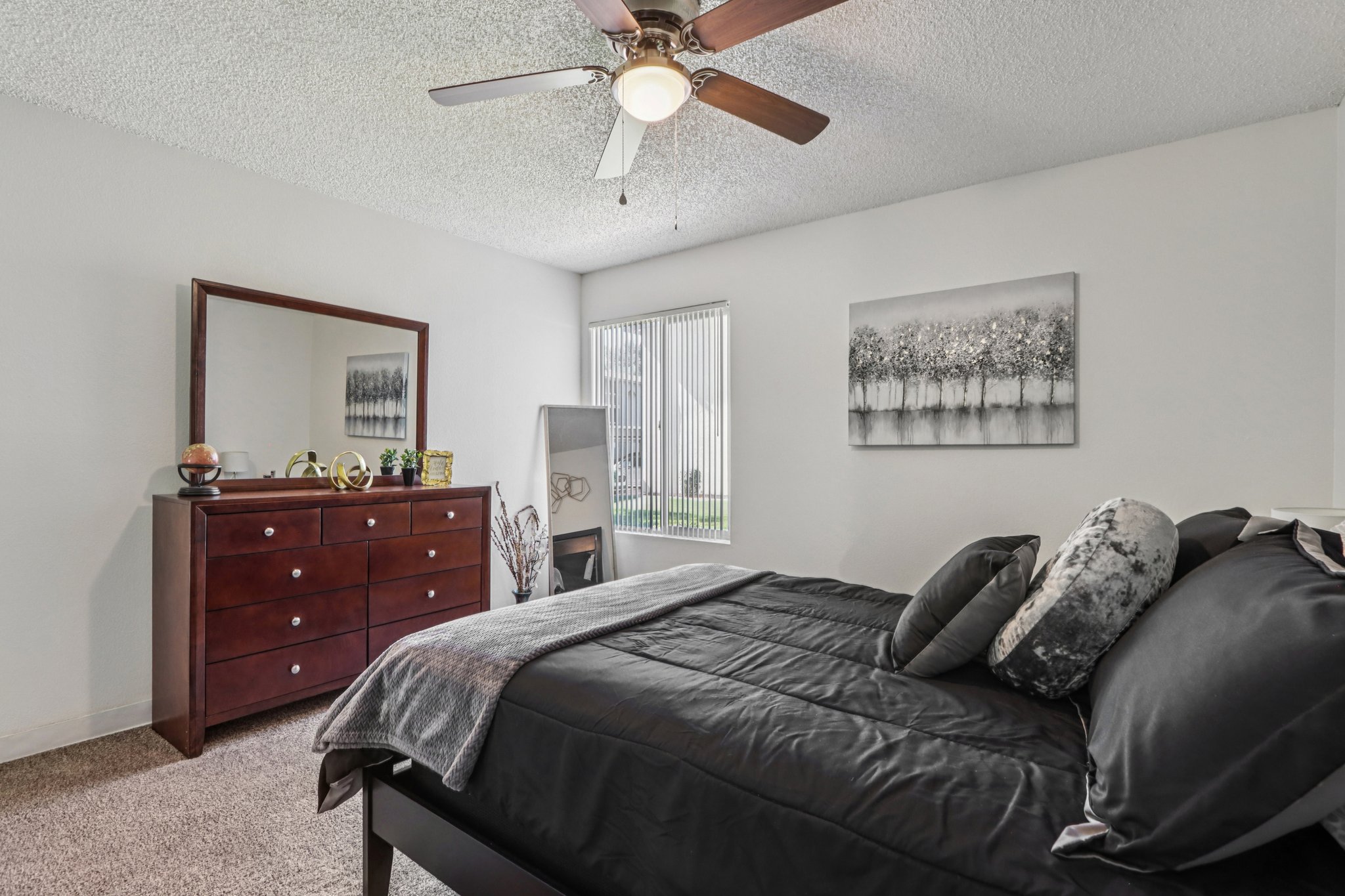 A cozy bedroom featuring a queen-sized bed with dark bedding, a wooden dresser with decorative items, a framed artwork on the wall, and a window with vertical blinds. A ceiling fan provides ventilation in the room, which has light-colored walls and carpeted flooring.