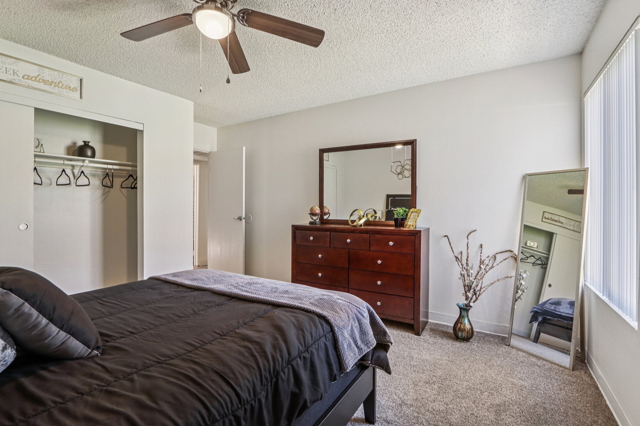 A cozy bedroom featuring a dark bedspread on a neatly made bed, a wooden dresser with decorative items, a large mirror, and a closet with hangers visible. Natural light filters in through a window with vertical blinds, creating a warm and inviting atmosphere.