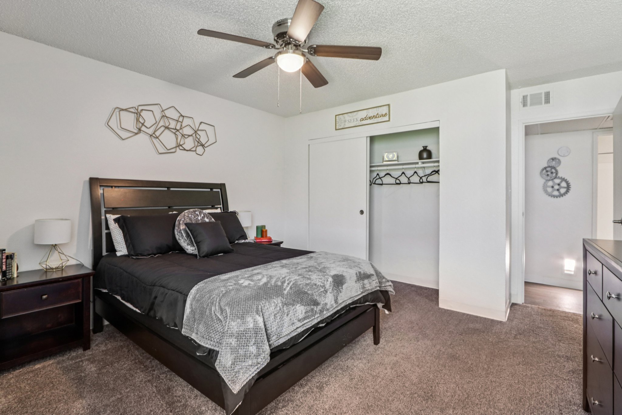 A modern bedroom featuring a black bed with gray and black bedding, two bedside tables with lamps, and a decorative wall piece above the bed. There is a closet with hangers and a door leading to another room. The walls are painted white, and the flooring is carpeted in a light shade. A ceiling fan is visible.