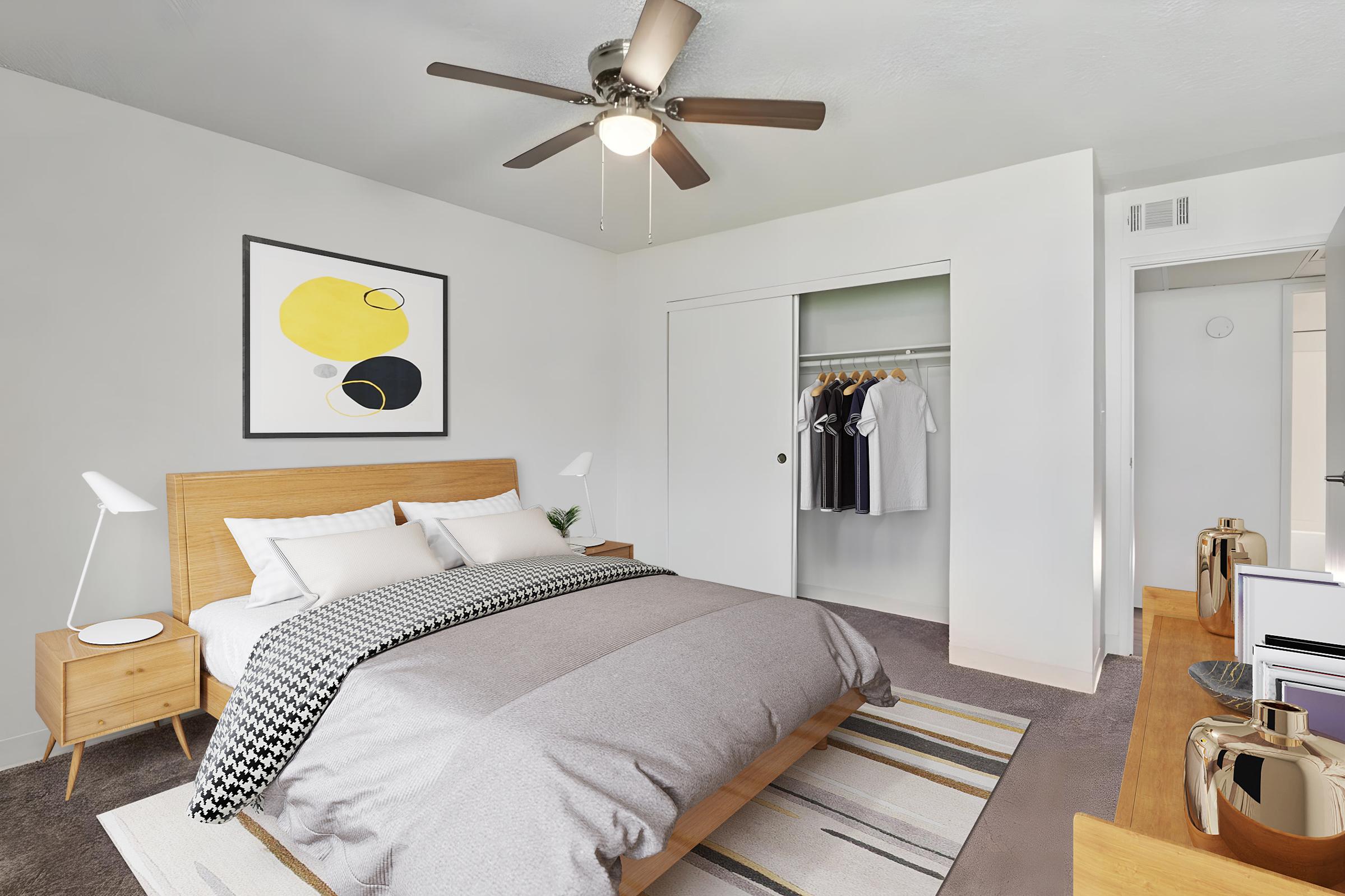 A modern bedroom featuring a wooden bed with gray bedding, two bedside lamps, and a decorative wall art piece in yellow and black. It has a closet with hanging clothes, a ceiling fan, and a light-colored rug. The walls are painted white, creating a bright and minimalist aesthetic.