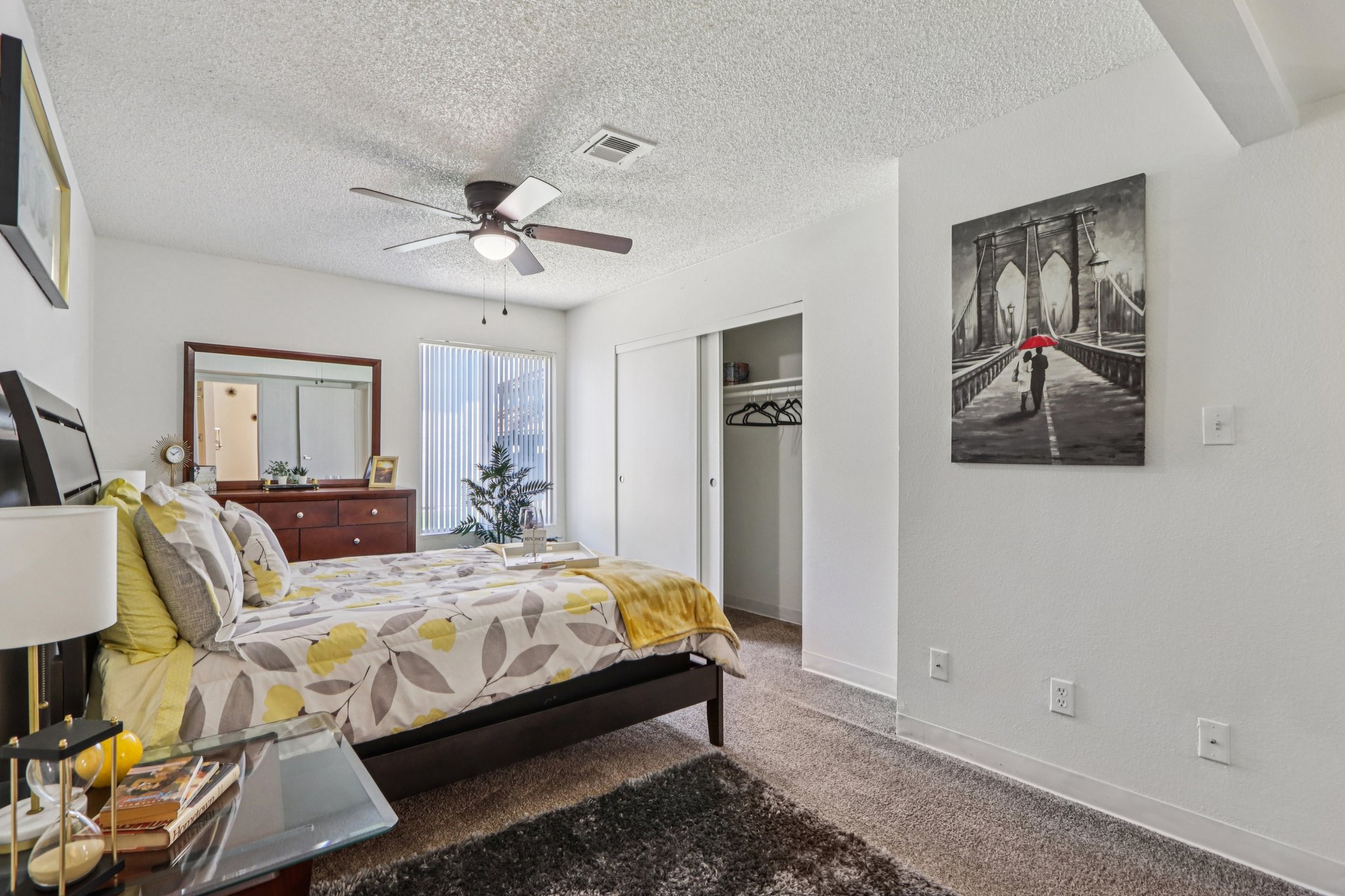 A cozy bedroom featuring a queen-sized bed with a patterned yellow and gray comforter, a ceiling fan, and a large window with light filtering through. A dresser and a mirror are visible, along with a closet and a piece of art depicting a bridge. The space has carpeted flooring and a warm, inviting atmosphere.