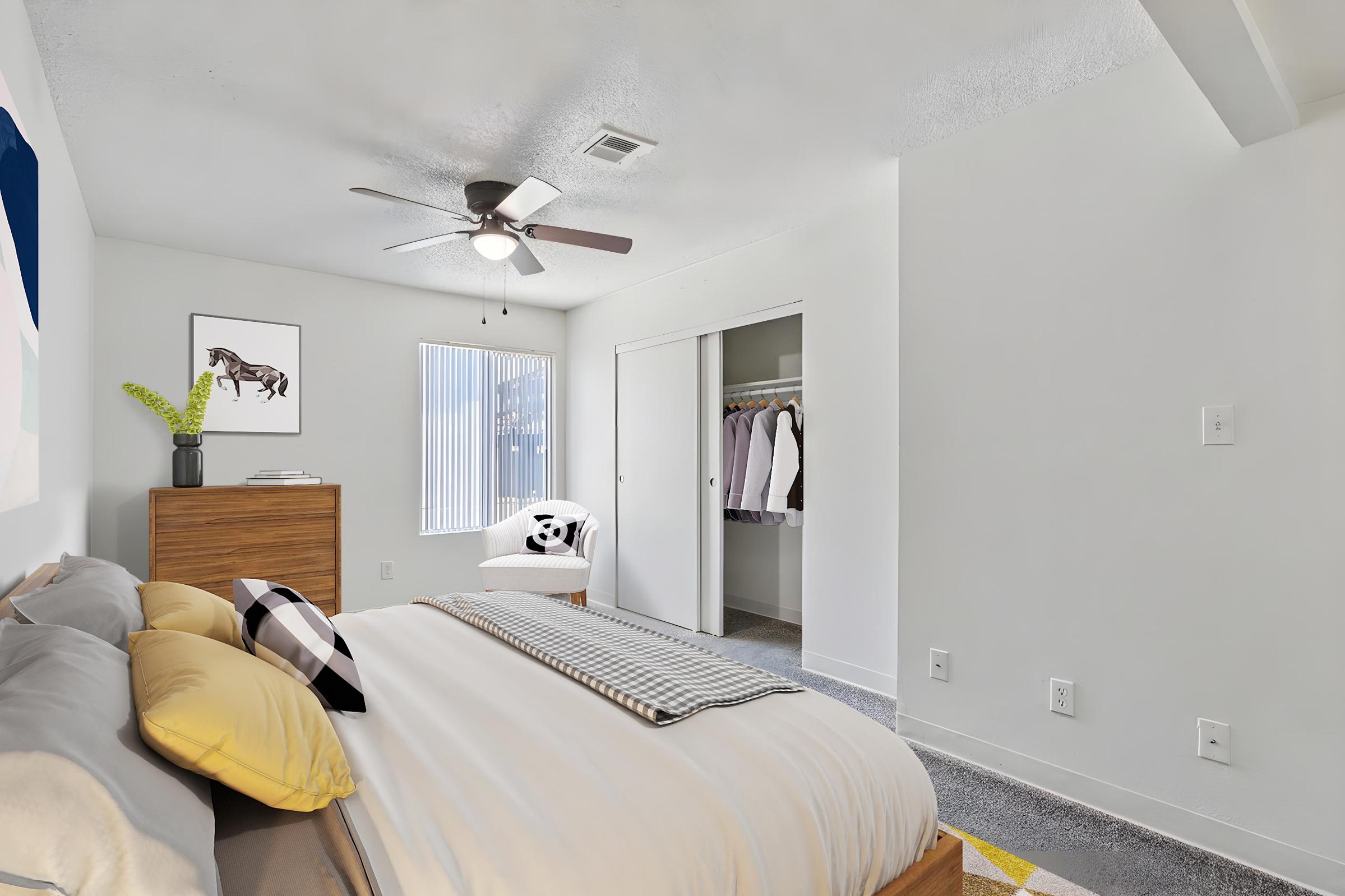 A cozy bedroom featuring a large bed with yellow and gray pillows, a modern wooden nightstand, and a ceiling fan. There is an artwork of a horse on the wall, and a chair with a panda-themed cushion next to a closet. Natural light filters through a window with blinds. 
