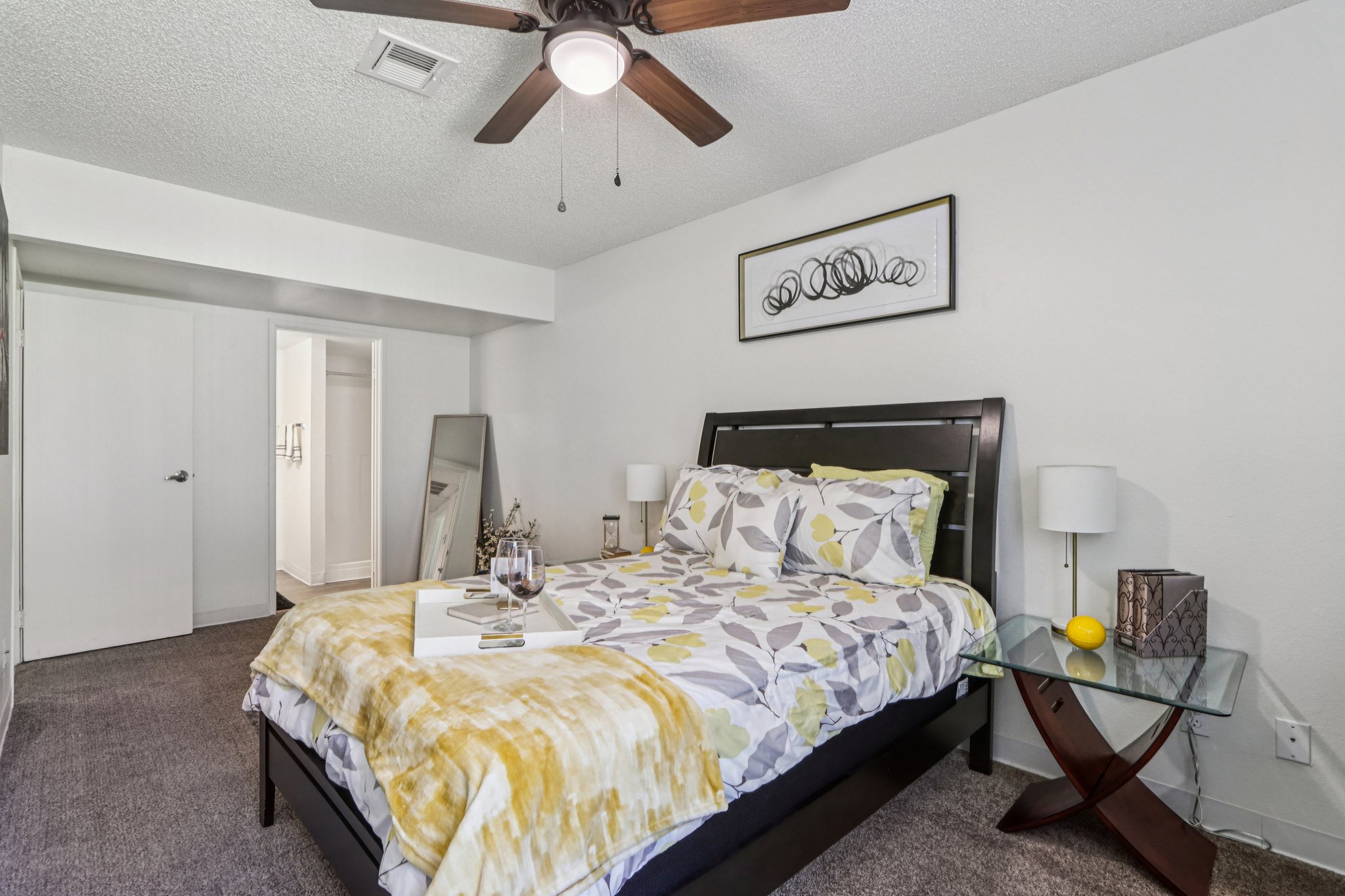 A cozy bedroom featuring a dark wooden bed with yellow and gray floral bedding, a decorative mirror, and a small glass nightstand with a glass of wine and a lemon. The room has light-colored walls, carpeted flooring, and a ceiling fan, adding to its inviting atmosphere. A bathroom entrance is visible in the background.