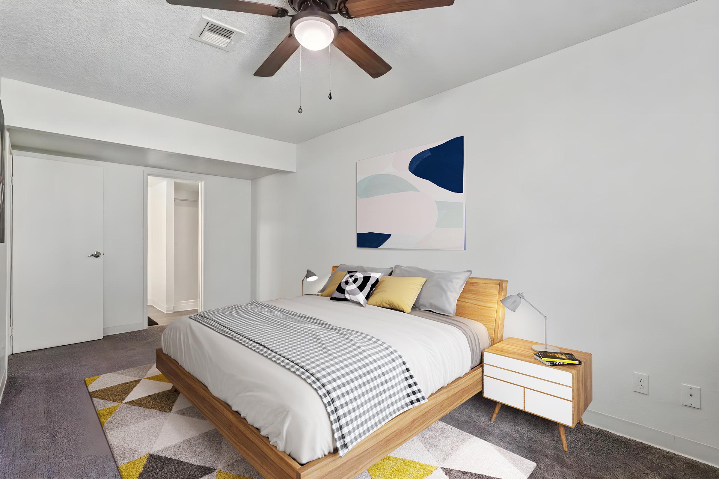 A modern bedroom featuring a wooden bed with gray bedding and decorative pillows. There is a light-colored rug with geometric patterns on the floor, a nightstand with a lamp, and a ceiling fan. A doorway leads to a closet, and an abstract painting hangs on the wall. The walls are painted white, creating a bright atmosphere.