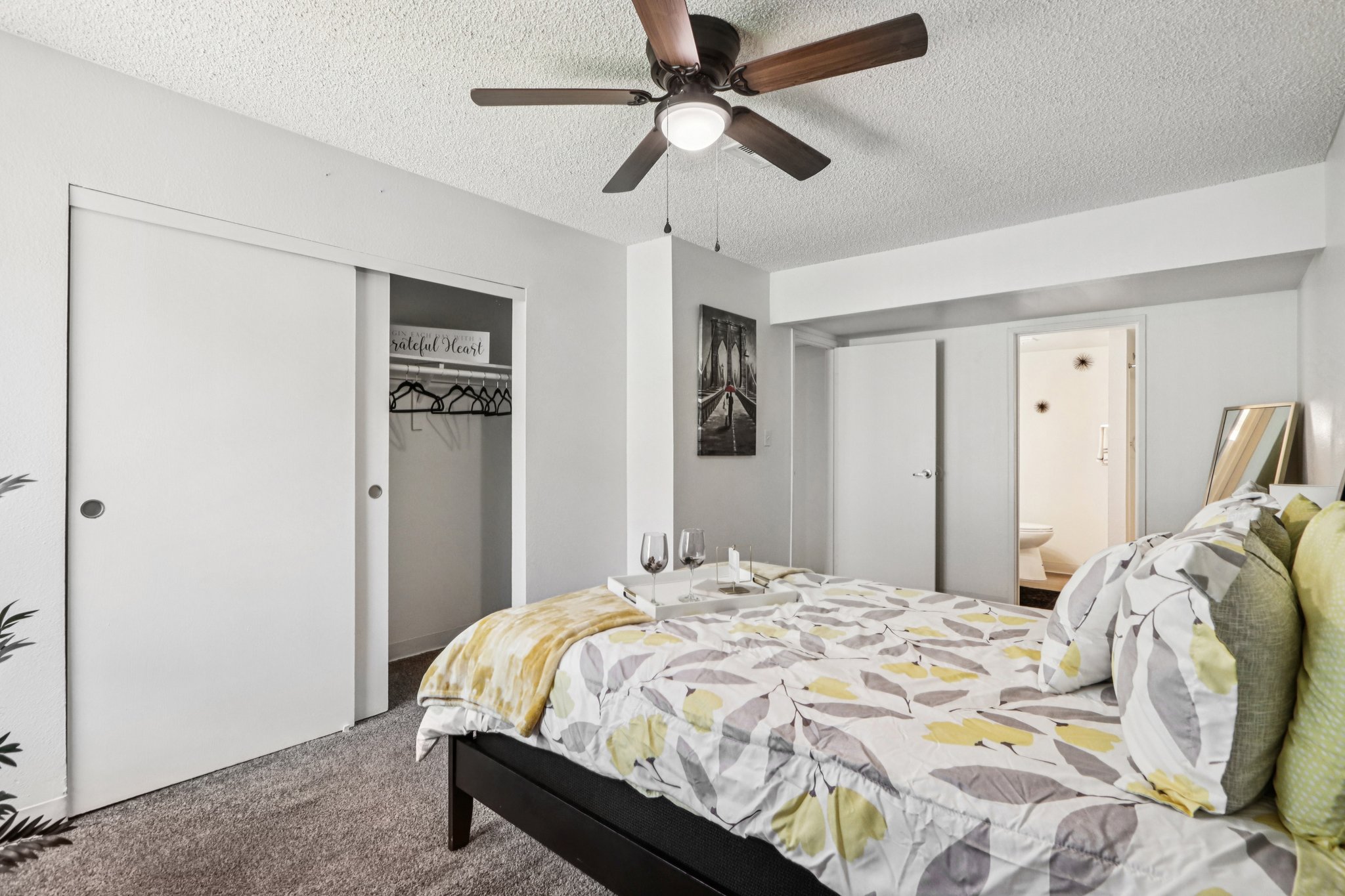 A cozy bedroom featuring a queen-sized bed with a floral comforter, two wine glasses on a tray, a ceiling fan, and a closet. The walls are painted light color, and there is a piece of artwork depicting a serene landscape. The room has carpet flooring and opens to a bathroom area.