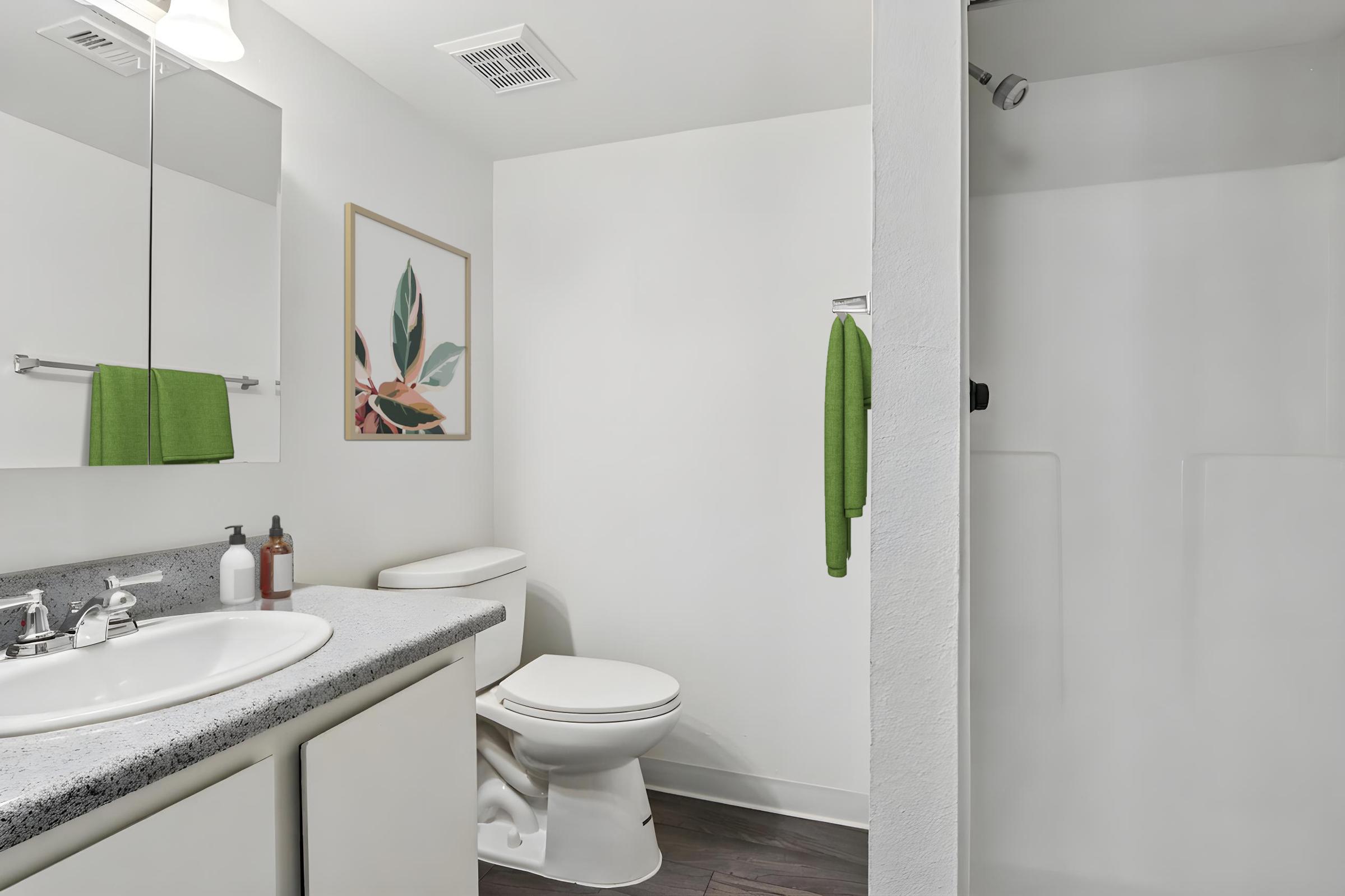 A modern bathroom featuring a white shower, a toilet, and a gray countertop with sink and mirror. There are green towels hanging on the towel rack, and a framed artwork with leaves on the wall. The design is clean and minimalistic, with light-colored walls and dark wood flooring.