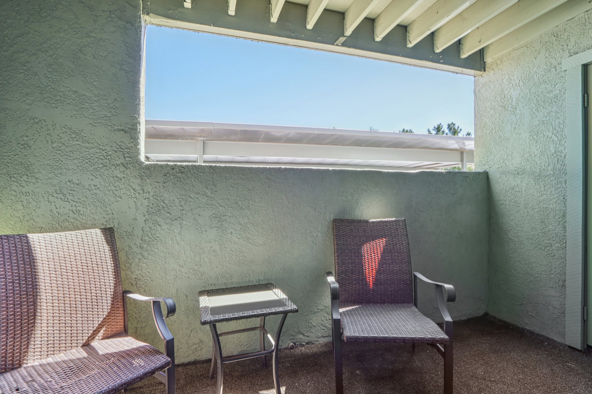 A small balcony with two wicker chairs and a glass table overlooking a bright sky. The walls are painted a light green, and there’s a slatted ceiling above. A view of a white canopy or awning is visible through the opening.