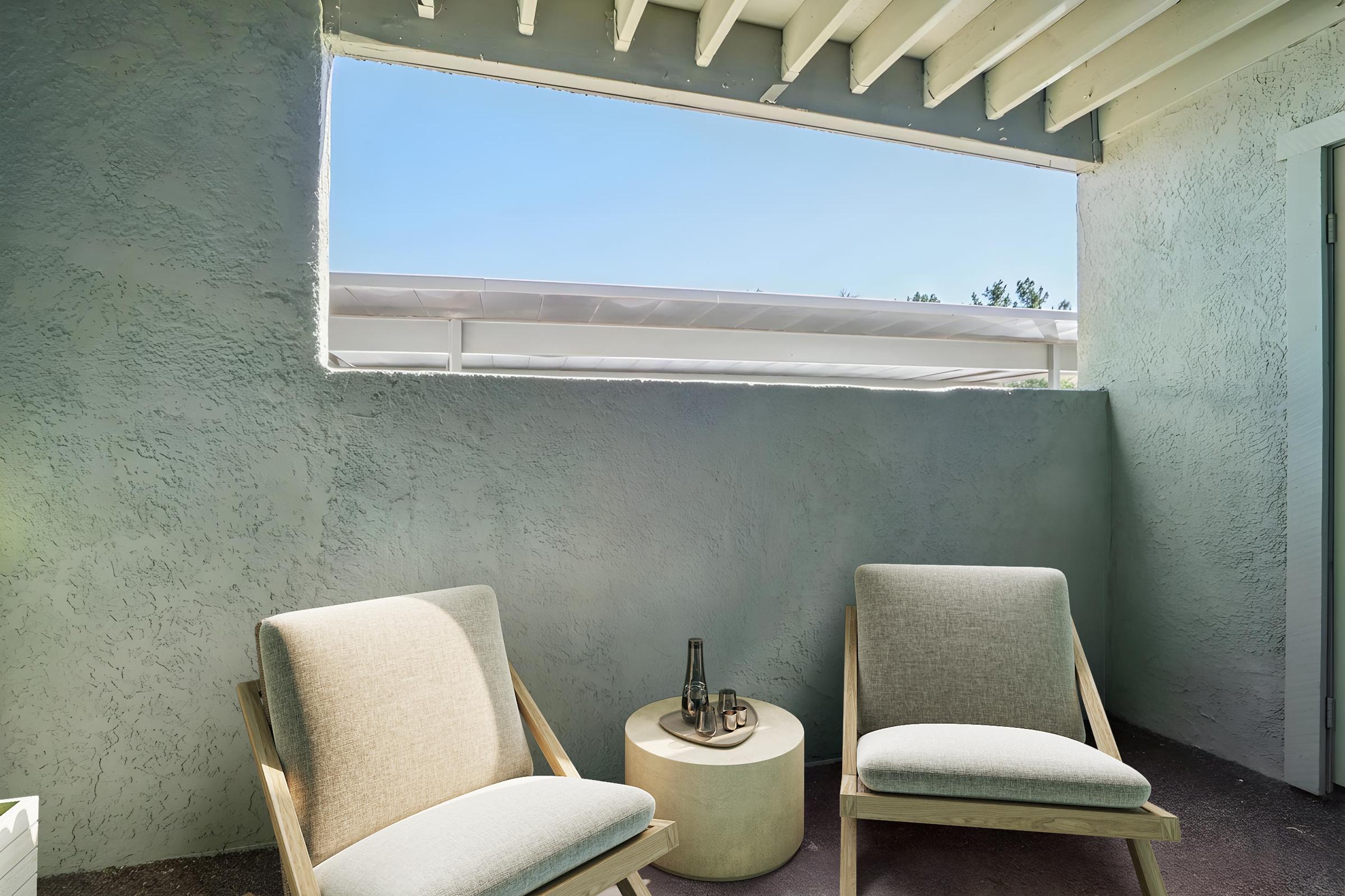 A cozy outdoor seating area featuring two light gray chairs and a round side table, set against a textured light gray wall. Above, a window lets in natural light, revealing a clear blue sky. The overall atmosphere feels serene and inviting.