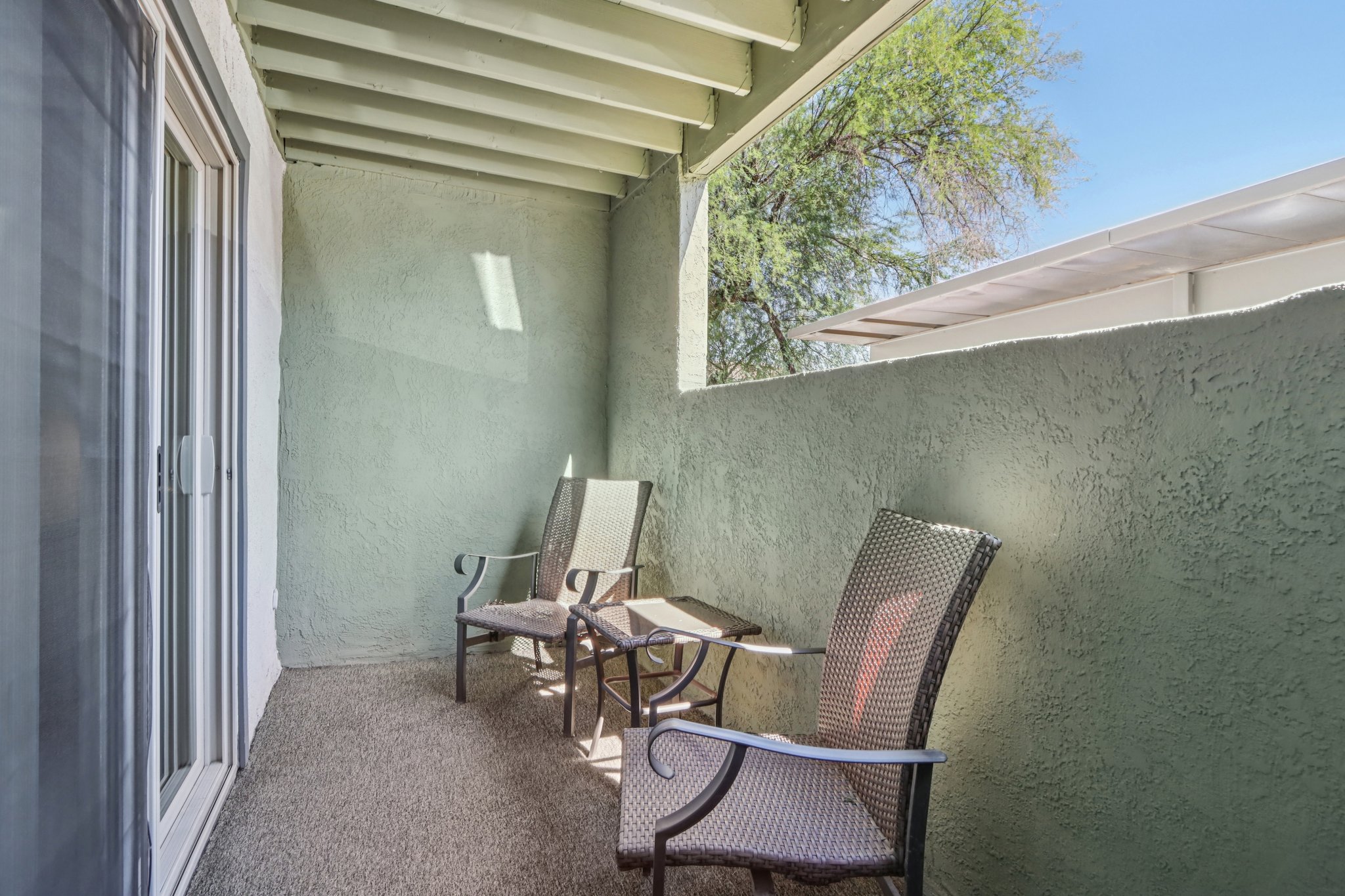 A small balcony with two chairs and a side table, surrounded by light green walls. The space is well-lit, with sunlight casting shadows on the floor, and features a textured wall finish. In the background, there are hints of trees and another structure visible.