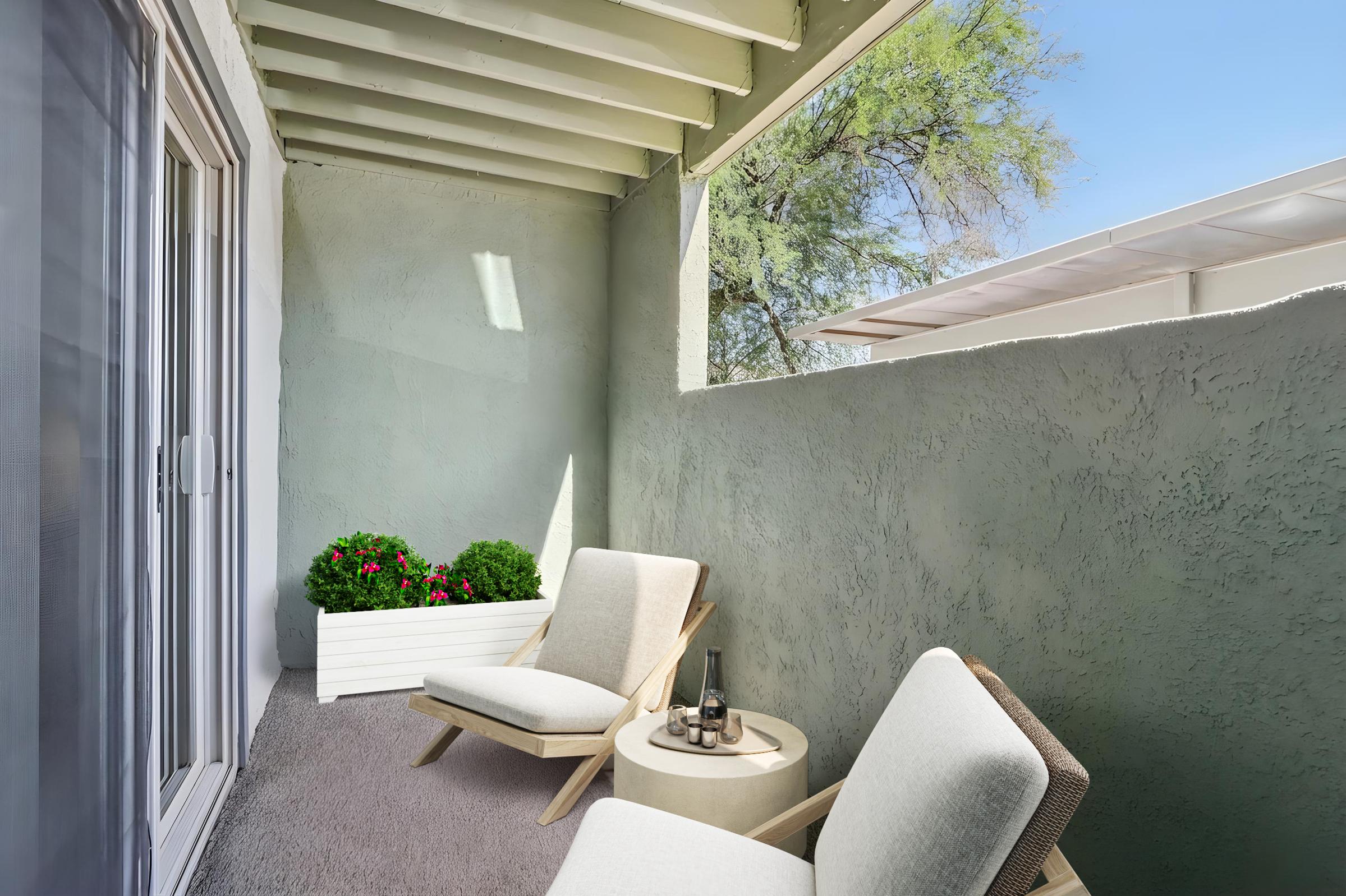 A cozy balcony featuring two wooden lounge chairs and a small round table. A white planter with vibrant flowers adds a touch of greenery. The walls are painted a soft gray, and there is a clear blue sky above, creating a bright and inviting outdoor space.