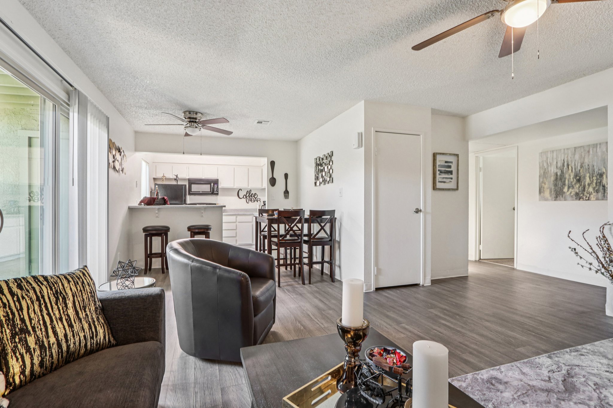 Interior view of a modern living space featuring a cozy seating area with a black chair and a decorative couch. In the background, a kitchen with a bar and stools is visible, alongside a dining area. Natural light floods the room through large windows, highlighting the contemporary decor and wooden flooring.