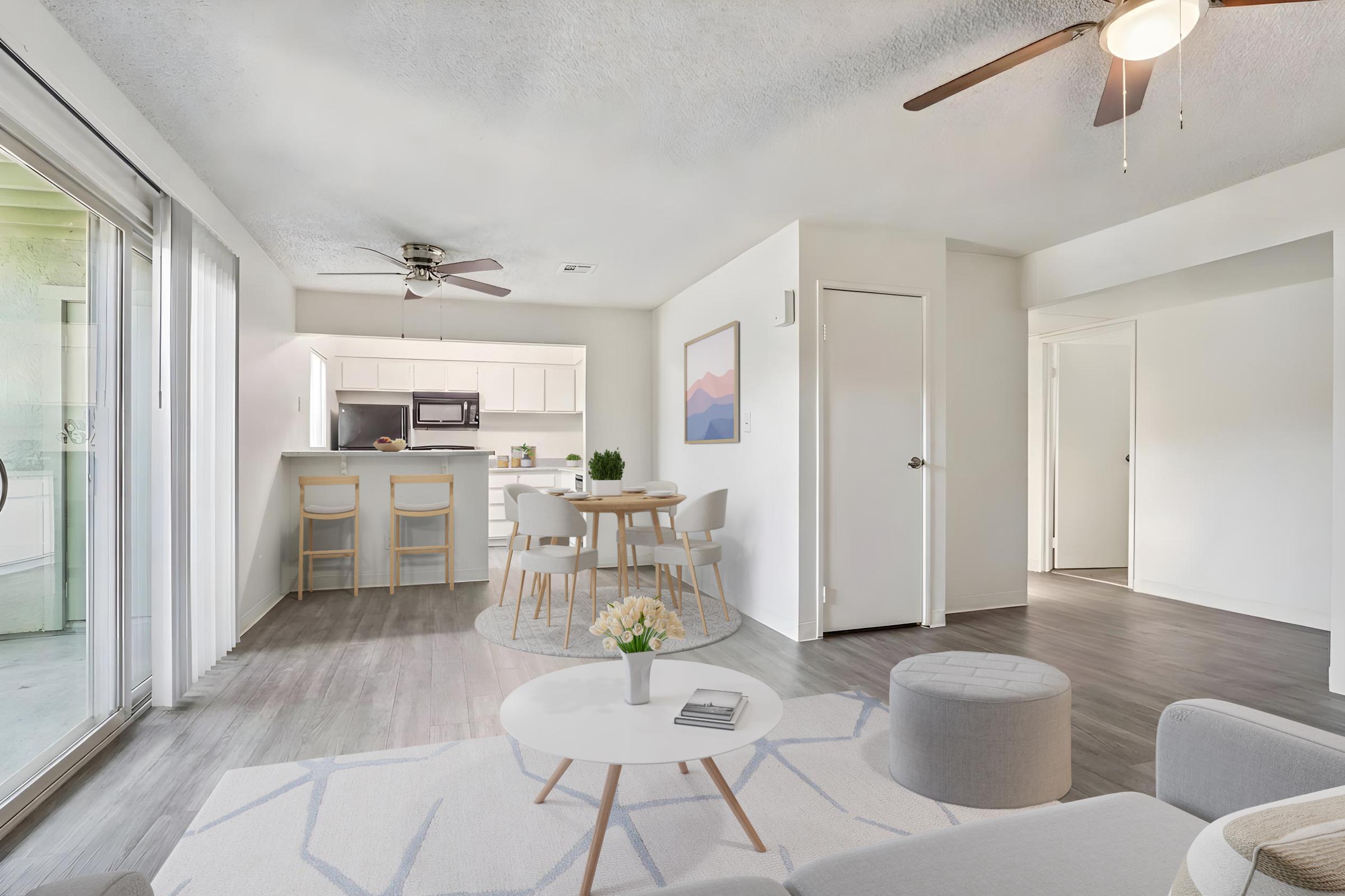 Bright and modern living space showcasing a cozy living area with a light rug and a small table. The dining area features a round table with four chairs and a kitchen visible in the background. Natural light floods the room through sliding glass doors, enhancing the open and inviting atmosphere.