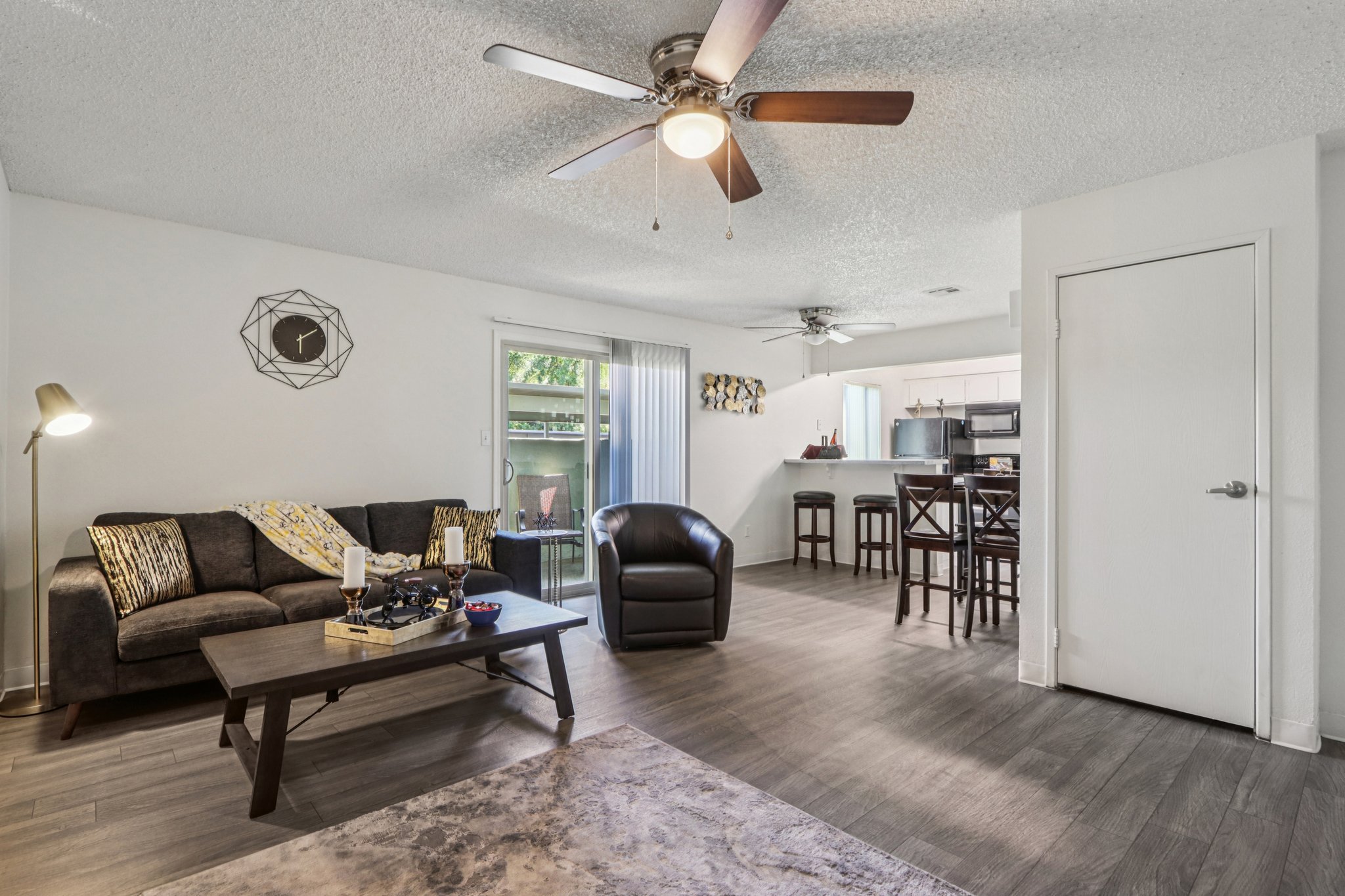 Living room featuring a dark sofa with yellow and gray accent pillows, a wooden coffee table with decorative items, a brown armchair, and ceiling fans. In the background, there is a kitchen area visible through sliding glass doors, with a modern dining setup and light-colored walls.