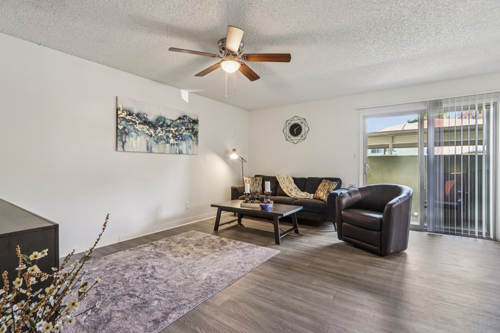 A cozy living room featuring a dark leather armchair, a coffee table with decorative items, and a beige area rug. There is a ceiling fan, a modern wall art piece, and sliding glass doors leading to an outdoor area. The light-colored walls create a bright, inviting atmosphere.