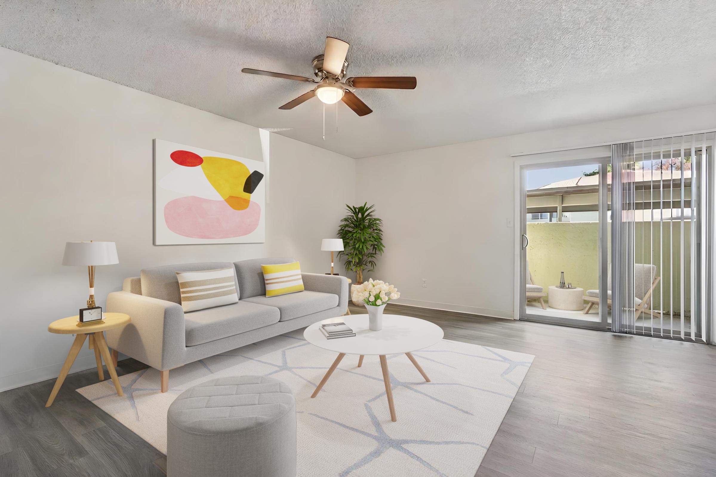 Modern living room featuring a light gray sofa with yellow and white striped cushions, a round coffee table on a geometric-patterned rug, and two lamps on side tables. A ceiling fan hangs above, and large sliding glass doors lead to a balcony with outdoor seating. The walls are painted in neutral tones, enhancing the airy feel.