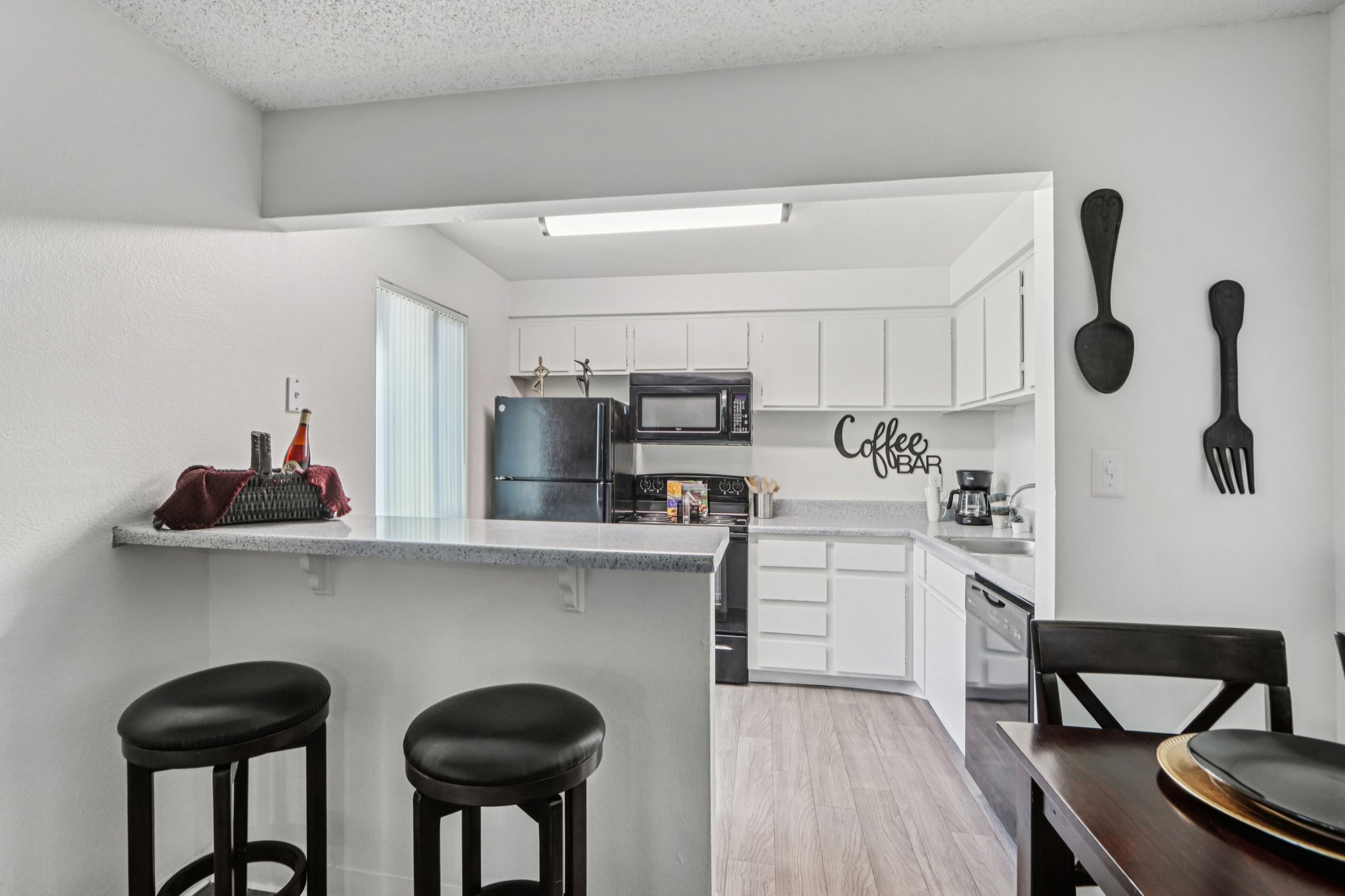 A modern kitchen featuring a breakfast bar with two black stools, a granite countertop, white cabinets, and stainless steel appliances. A "Coffee Bar" sign is displayed on the wall, enhancing the cozy atmosphere. Natural light filters in through a sliding glass door, giving a bright, inviting feel to the space.