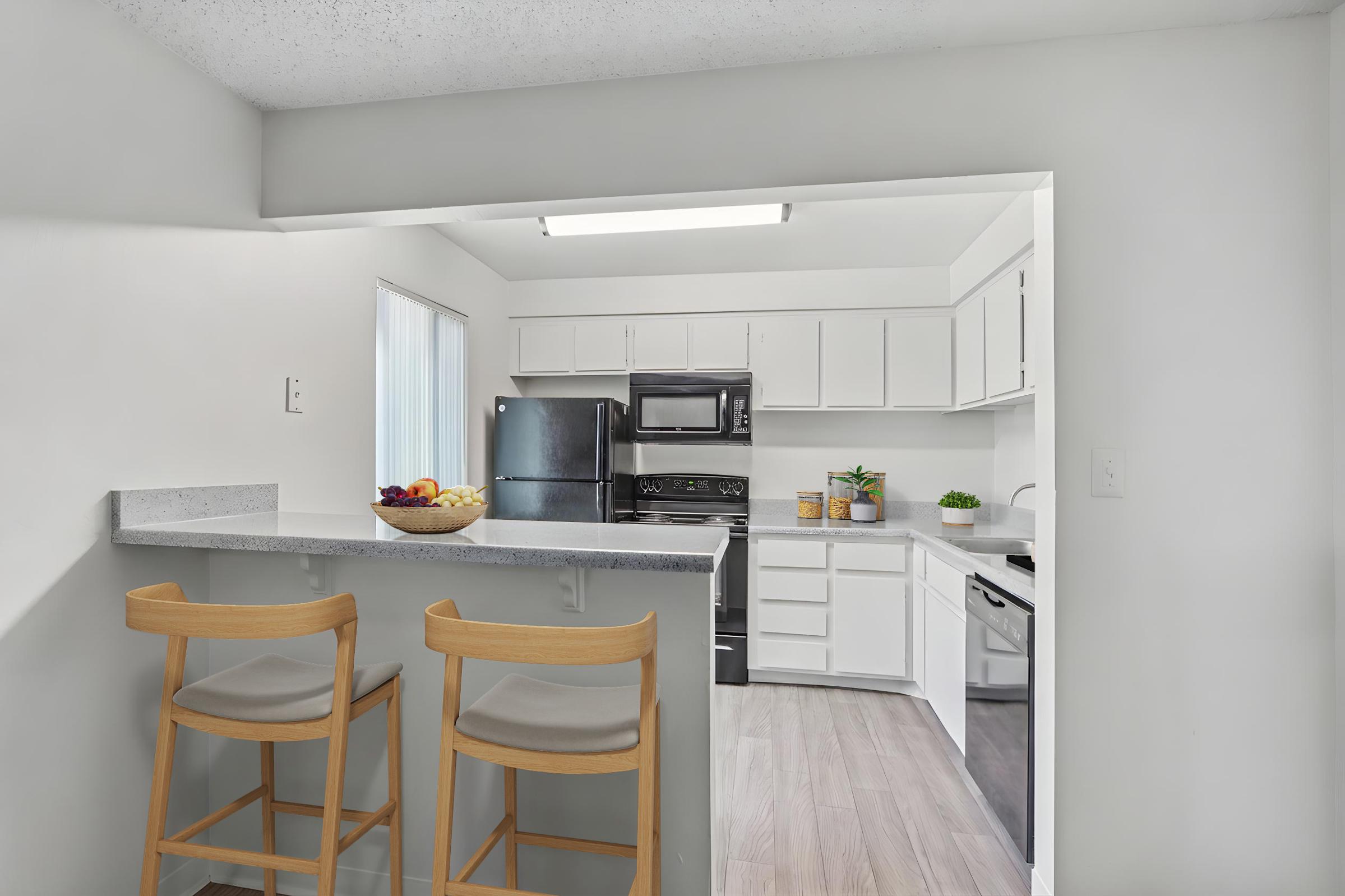 A modern kitchen with white cabinetry and stainless steel appliances. The area features a countertop with two wooden bar stools and a bowl of fruit. Large windows provide natural light, and potted plants add a touch of greenery to the space. The flooring is light-colored, enhancing the airy atmosphere.