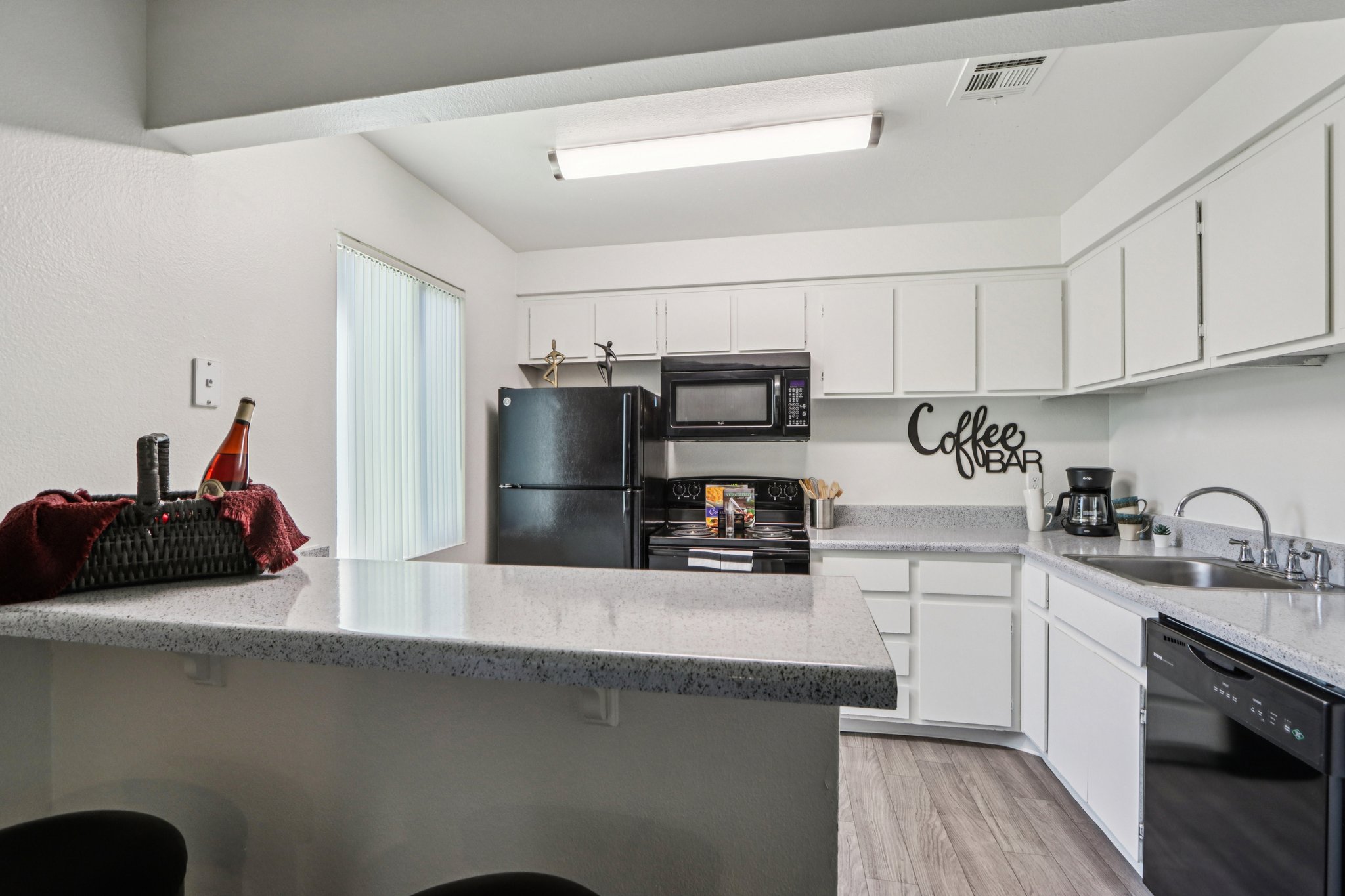 A modern kitchen featuring white cabinetry and a granite countertop. It includes a black refrigerator, microwave, and coffee maker. A basket with a bottle is on the bar area, and the wall has a decorative "Coffee Bar" sign. Soft natural light comes through a window with vertical blinds.