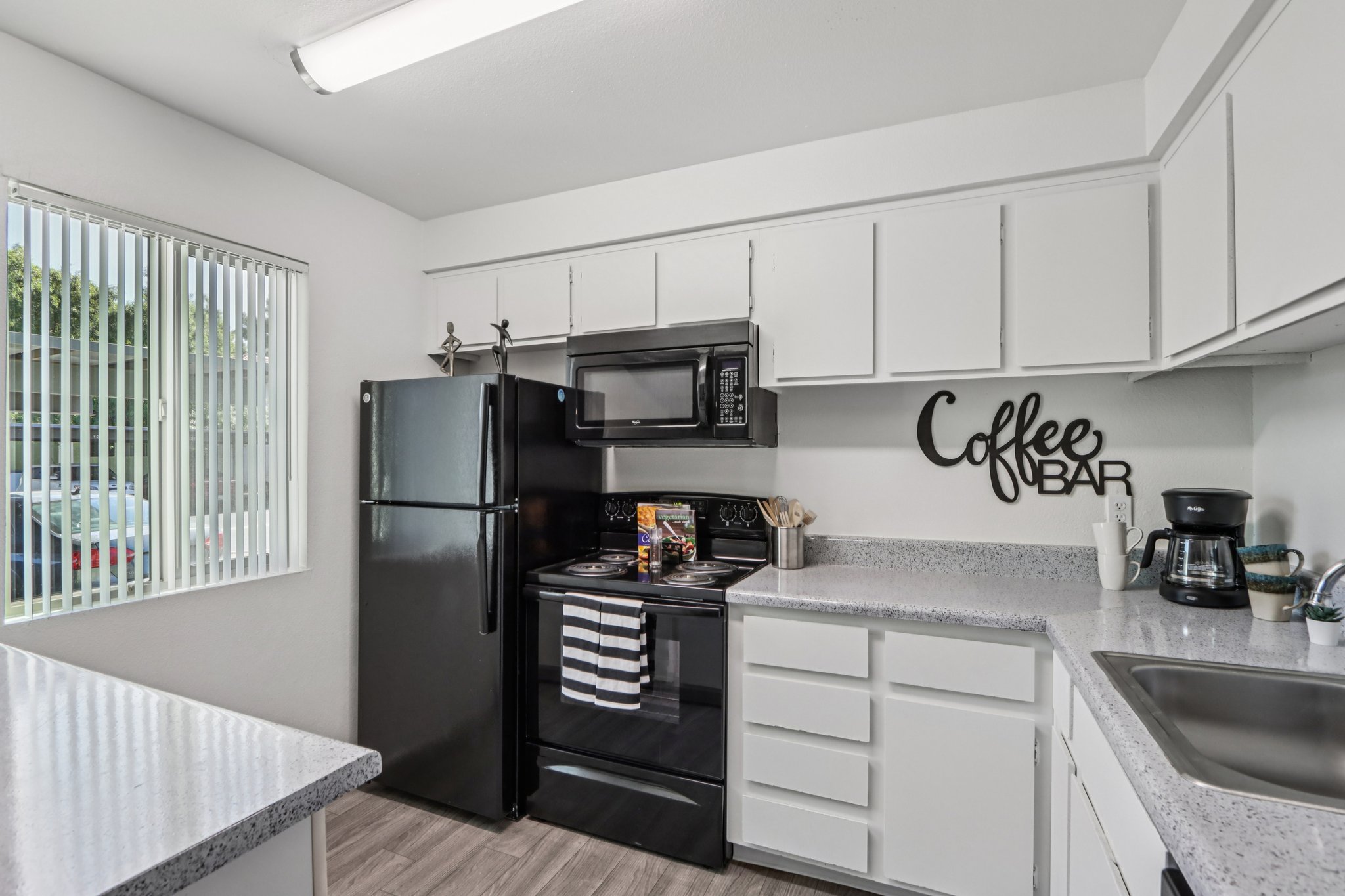 Modern kitchen featuring black appliances, including a refrigerator and microwave. A coffee maker is on the counter next to a "Coffee Bar" wall decoration. The space has white cabinetry and a light gray countertop, with natural light coming through a window adorned with vertical blinds.