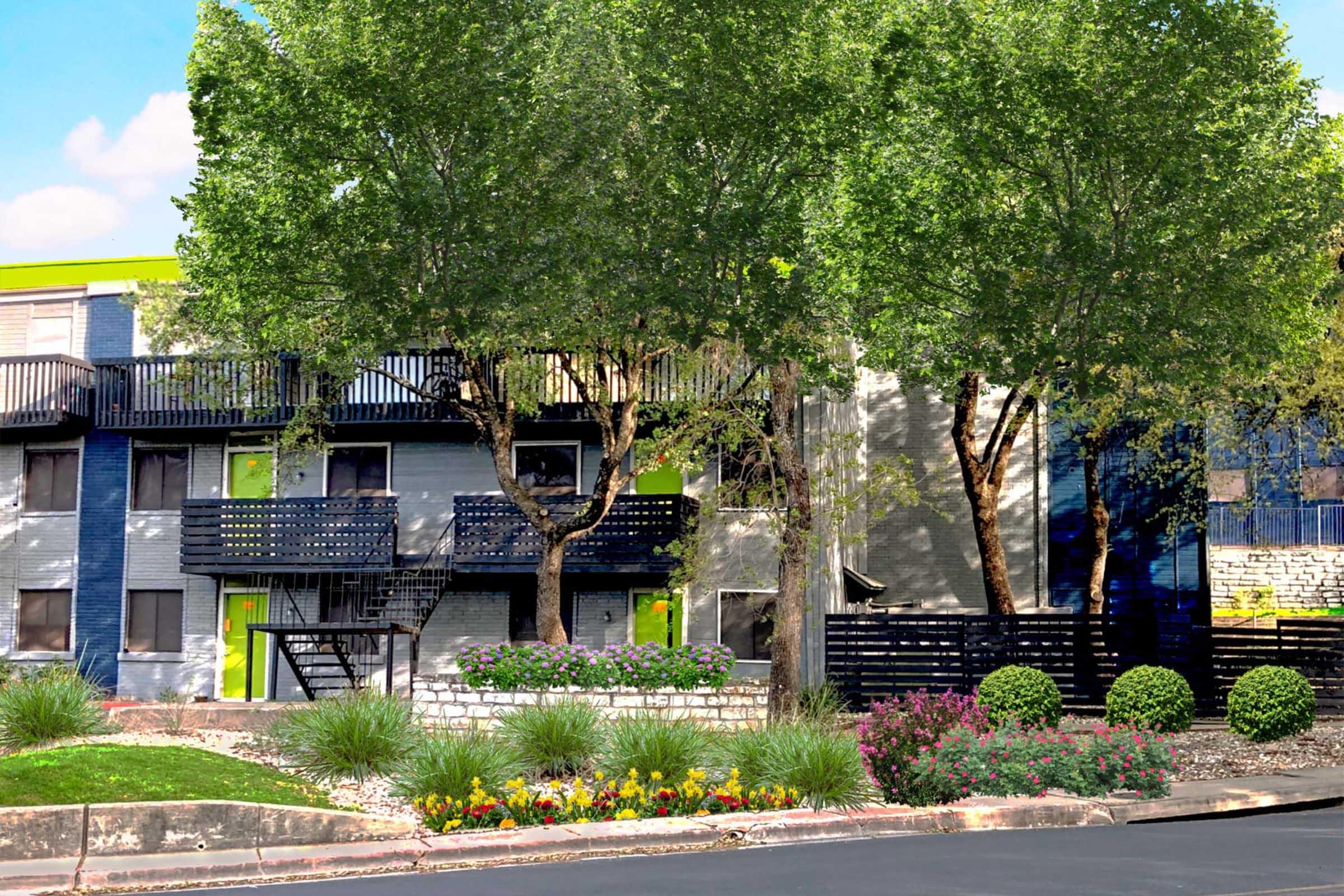 A view of a modern building with gray and green accents, featuring balconies and surrounded by lush trees and colorful flower beds. The landscaping includes well-maintained shrubs and a decorative stone wall, with a sunny blue sky in the background.