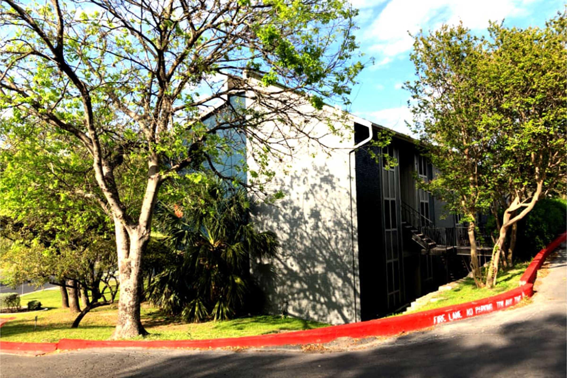 A two-story apartment building surrounded by trees and greenery. The building features a gray exterior with balconies. A red curb runs along the path in front of the building, and the sky is partly cloudy, indicating a bright day.