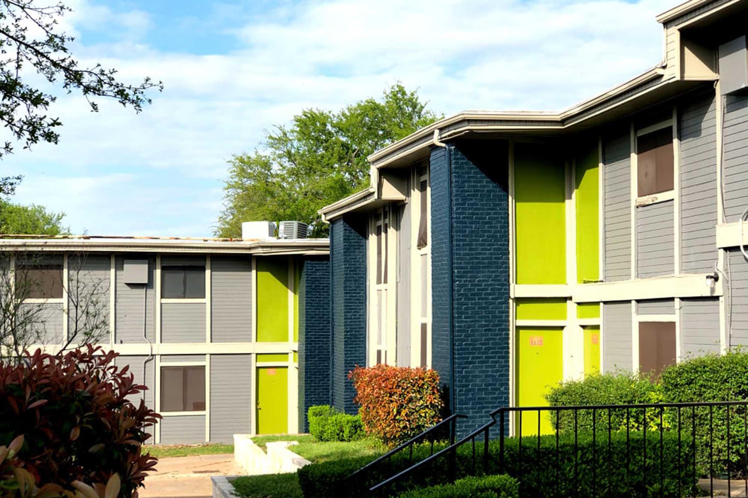 A modern apartment complex featuring gray and vibrant green exterior walls. The building has multiple units with large windows and a neatly landscaped area in front, including shrubs and a staircase leading to the entrance. The sky is partly cloudy, adding a bright ambiance to the scene.