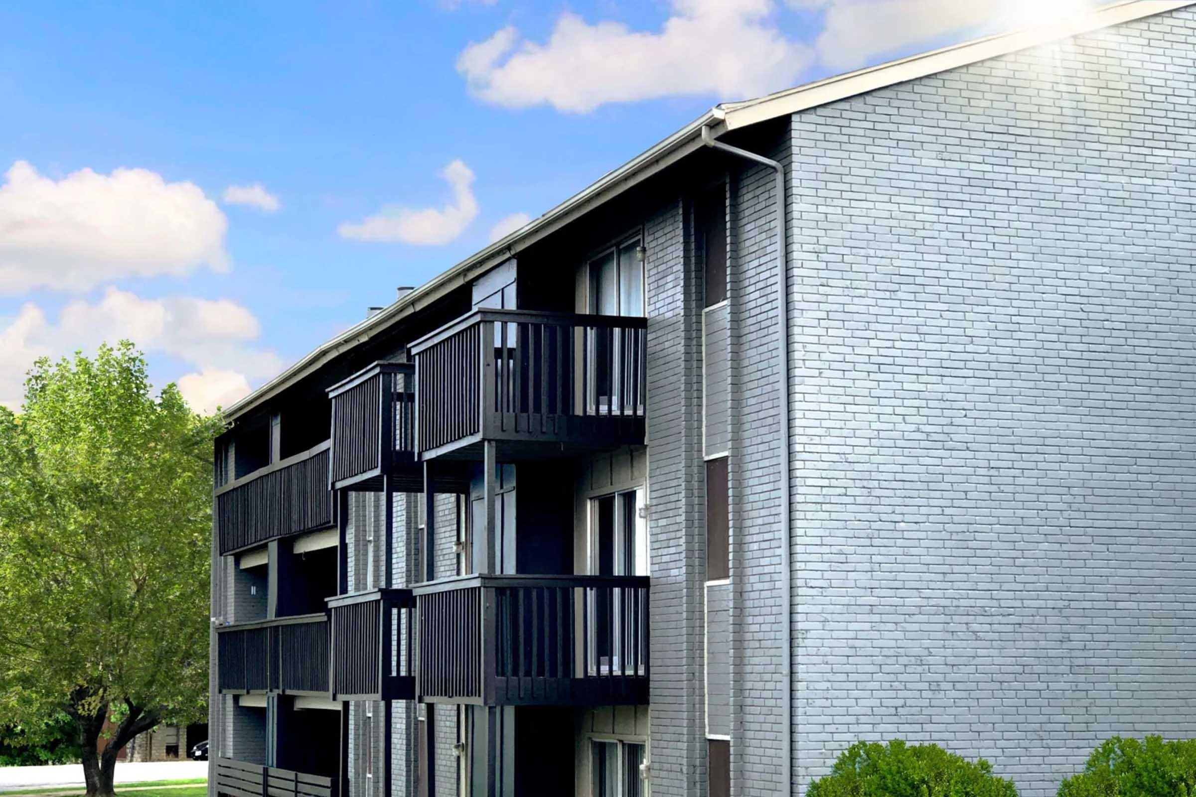 Three-story apartment building with a gray brick exterior and black balconies. The building is surrounded by greenery under a bright blue sky with scattered clouds.