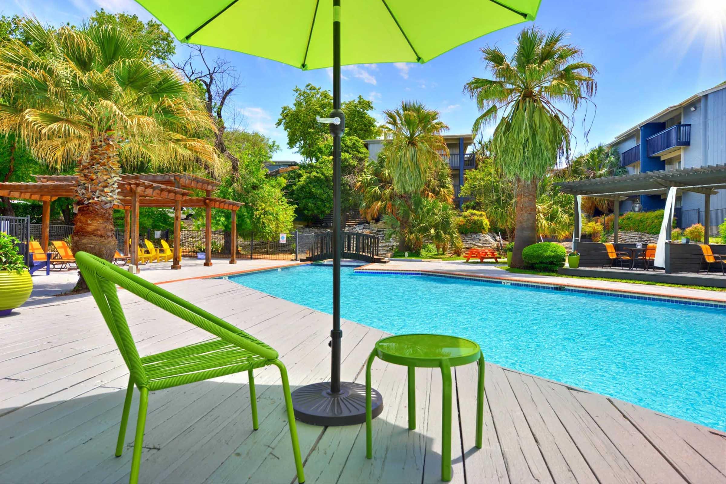 A vibrant outdoor scene featuring a bright green patio chair and small table under a matching umbrella. The setting includes a swimming pool surrounded by lush palm trees and colorful furniture, creating a relaxing atmosphere in a sunny environment.