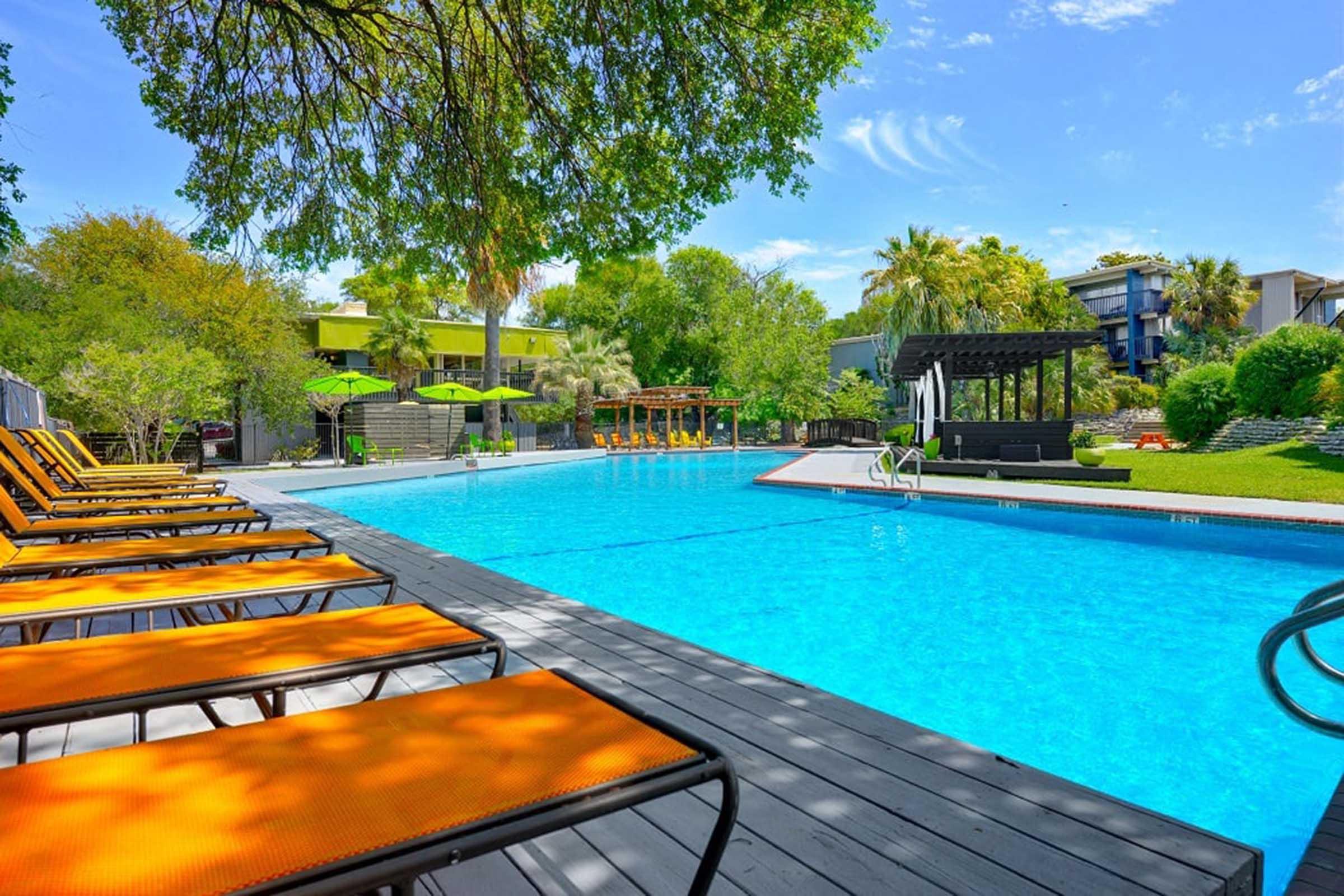 A bright swimming pool surrounded by lounge chairs with orange cushions. Lush green trees and landscaping are visible in the background, along with a shaded seating area and vibrant umbrellas. The clear blue sky complements the inviting atmosphere of the outdoor space.