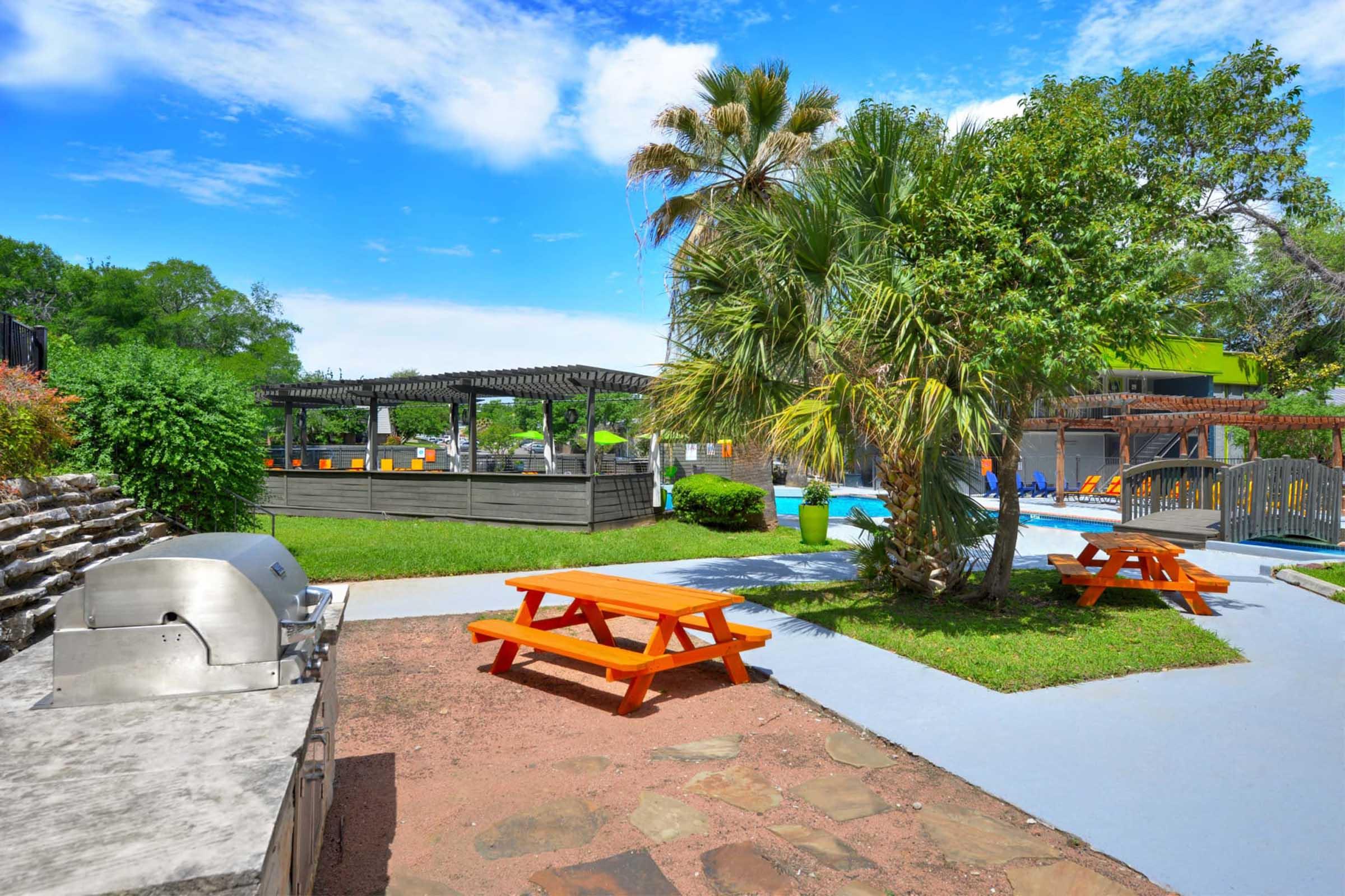 A bright outdoor space featuring a swimming pool surrounded by lounge chairs, picnic tables with orange tops, and a shaded area with pergolas. Lush greenery, including palm trees, adds to the relaxing atmosphere. A barbecue grill is visible in the foreground.