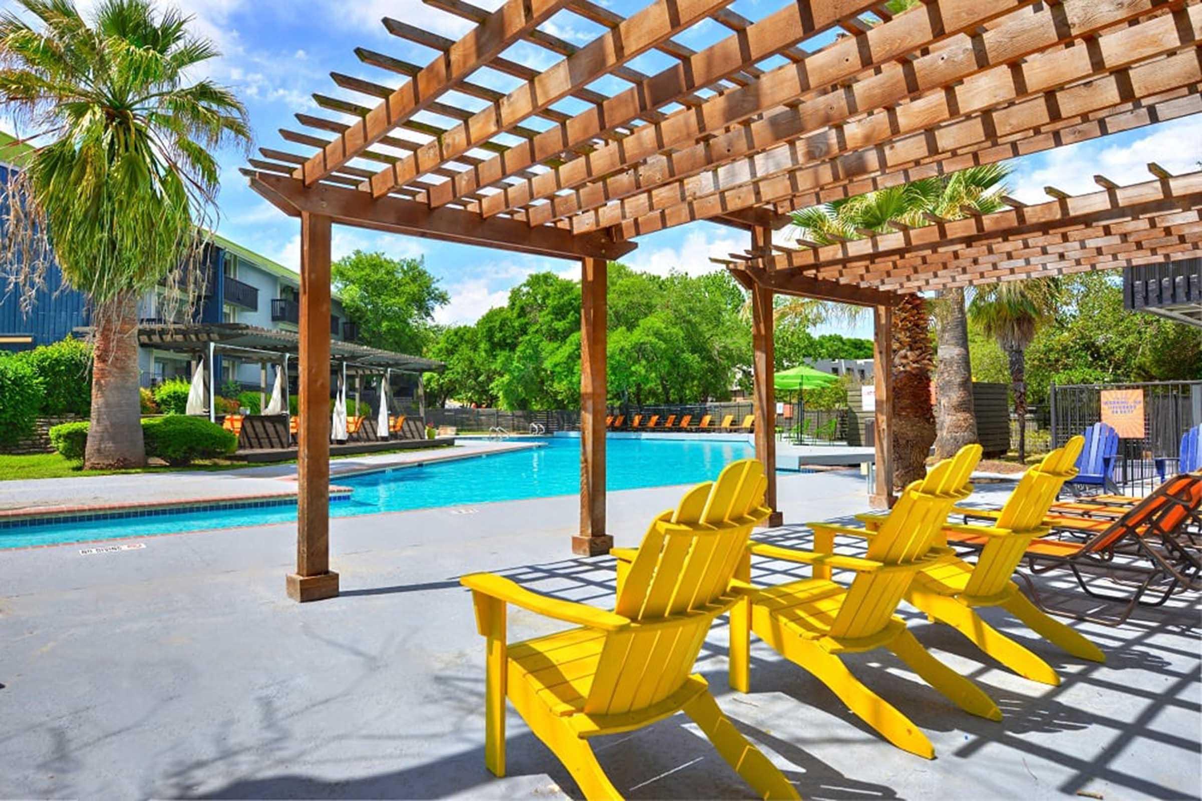 A sunny outdoor pool area featuring a blue swimming pool surrounded by yellow adirondack chairs under a wooden pergola. Lush greenery and palm trees are visible in the background, along with a few umbrellas. The scene is bright and inviting, perfect for relaxation.